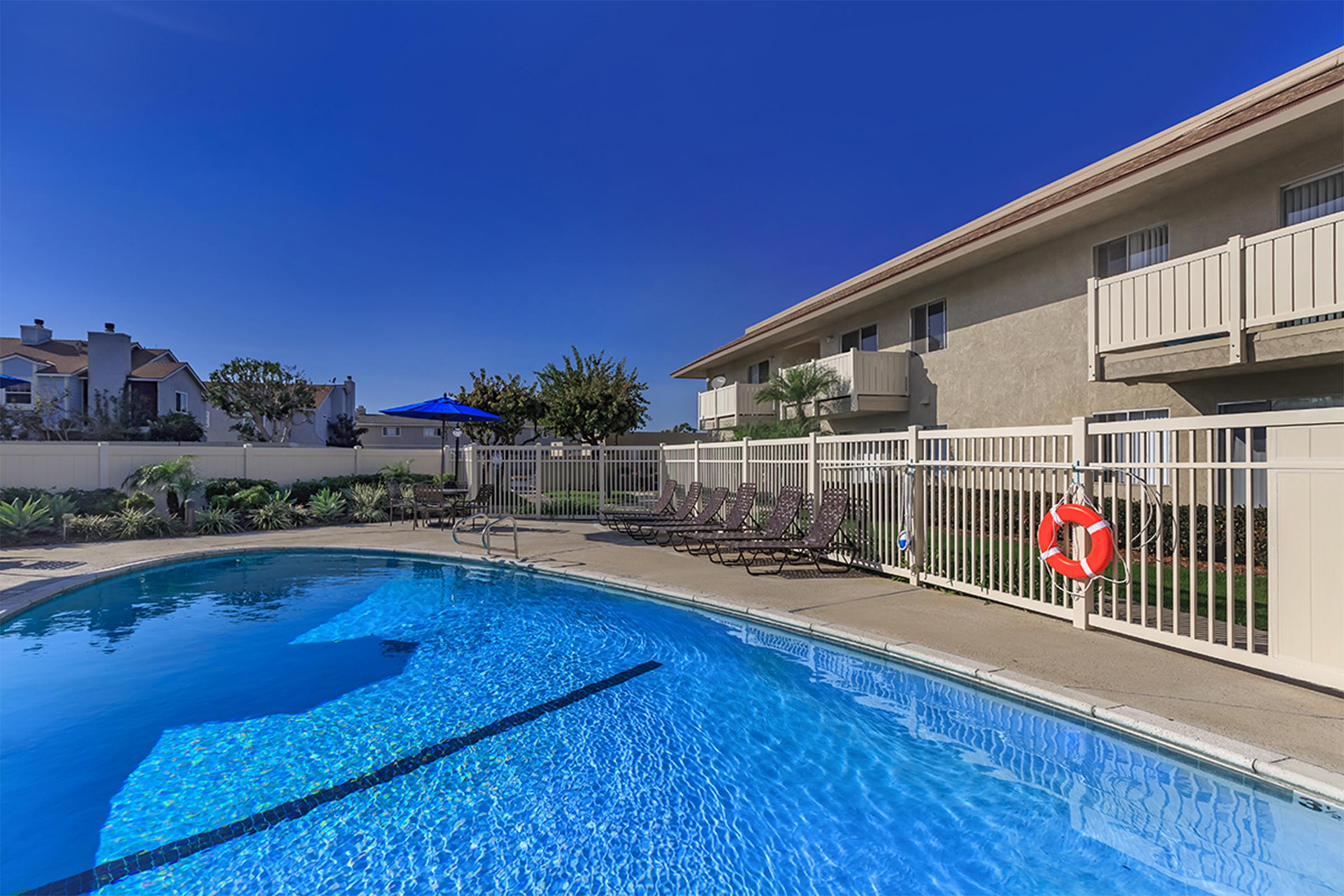 A clear swimming pool surrounded by a tan fence, with several lounge chairs lined up nearby. An umbrella provides shade over one area, and a bright blue sky is visible above. The setting features greenery and a residential building in the background, creating a relaxed outdoor atmosphere.