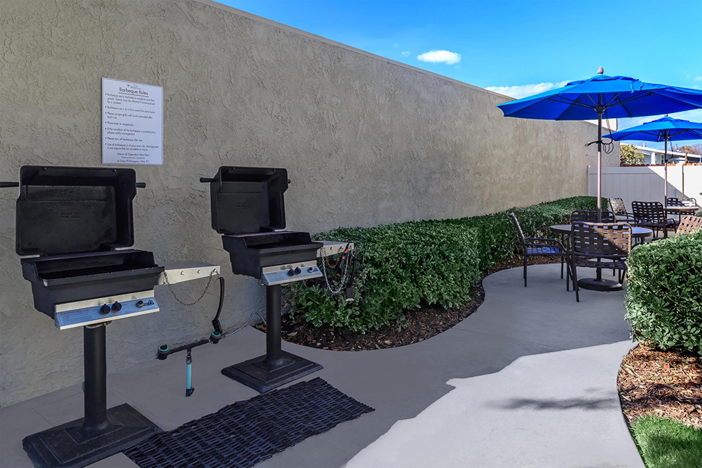 Two charcoal grills on stands next to a concrete path, with a menu sign on the wall nearby. In the background, there are blue umbrellas providing shade over outdoor seating with black chairs and tables, surrounded by neatly trimmed green shrubs under a clear blue sky.