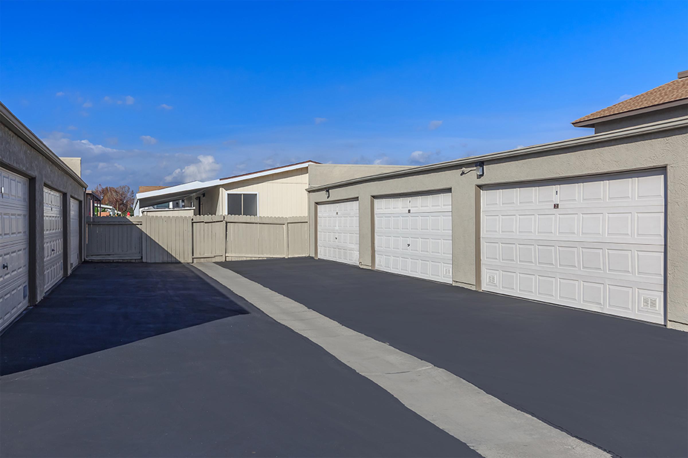 A clear view of a well-maintained parking lot featuring multiple closed garage doors on the right side. The pavement is freshly sealed in dark gray, and there is a wooden fence on the left. The sky is bright blue with a few clouds, suggesting a sunny day.