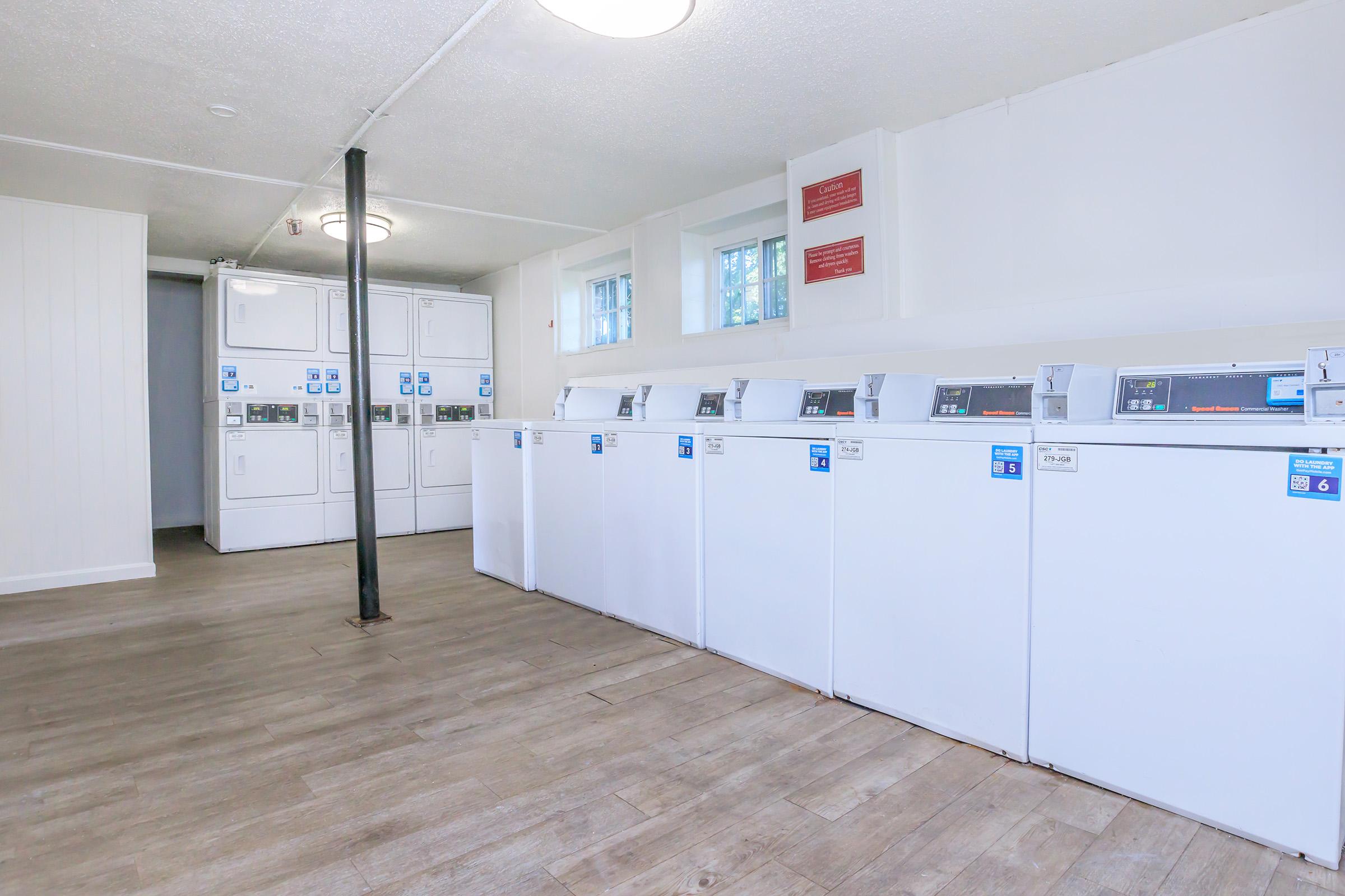 A bright and clean laundry room featuring multiple white washing machines and dryers lined up against a wall. The room has light-colored wooden flooring, overhead lighting, and windows that allow natural light to enter. Signs are displayed on the walls providing usage instructions.