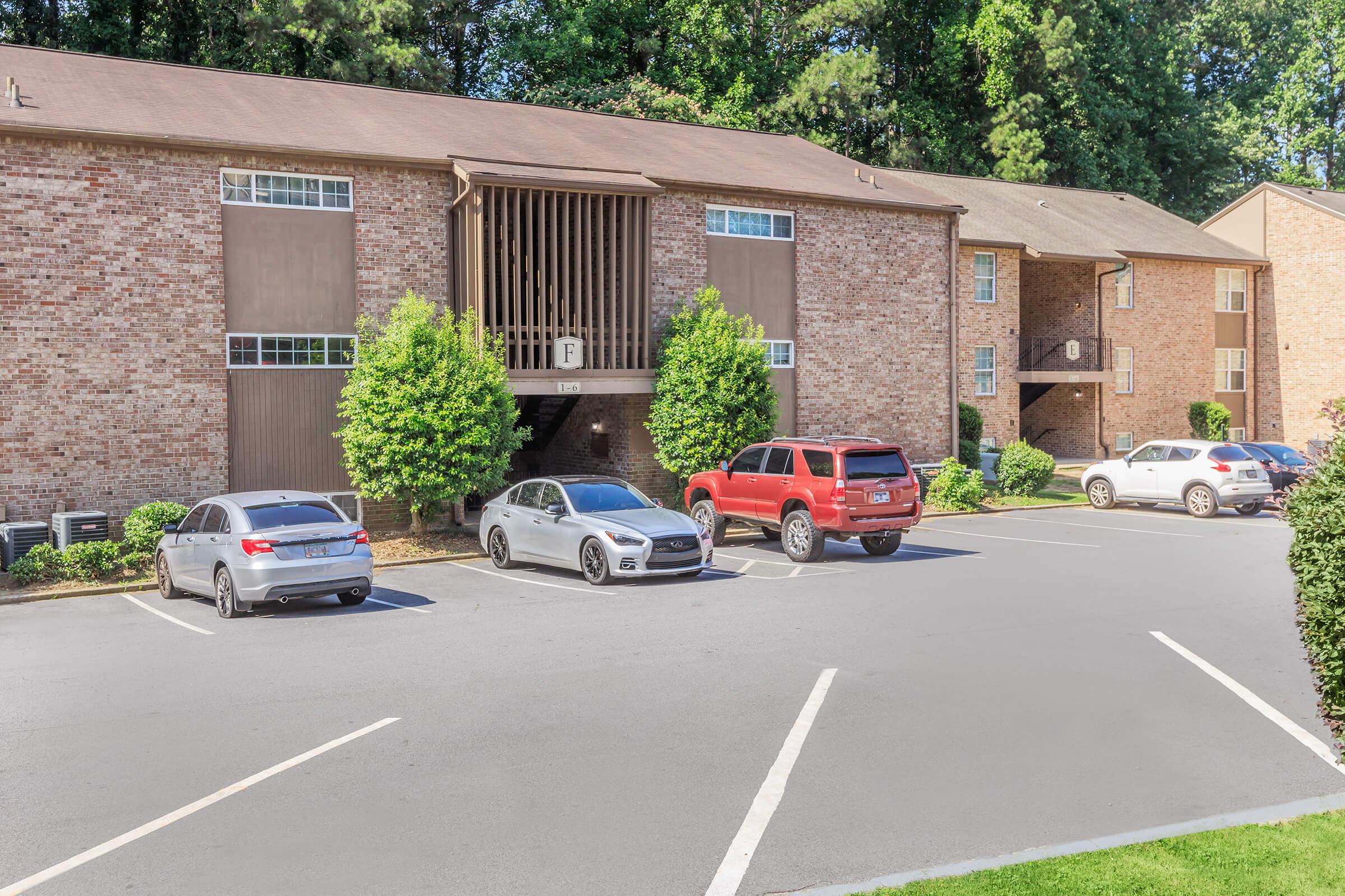 A view of an apartment complex with brick exteriors, featuring two buildings flanking a central entrance. In the foreground, there are four parked vehicles—two sedans and two SUVs—alongside well-maintained landscaping. The scene is bright and sunny, with trees in the background providing shade.