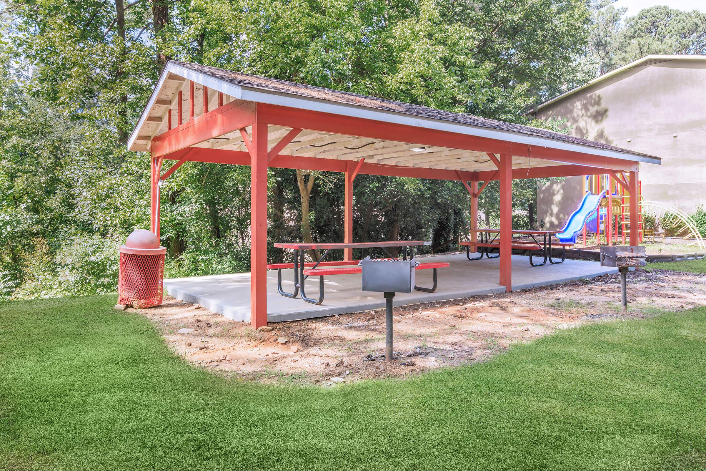 A covered picnic area with red wooden frames and picnic tables, situated in a green park. Nearby, there is a playground slide and a grill in front of the picnic area. Lush trees provide a natural backdrop, creating a serene outdoor space.