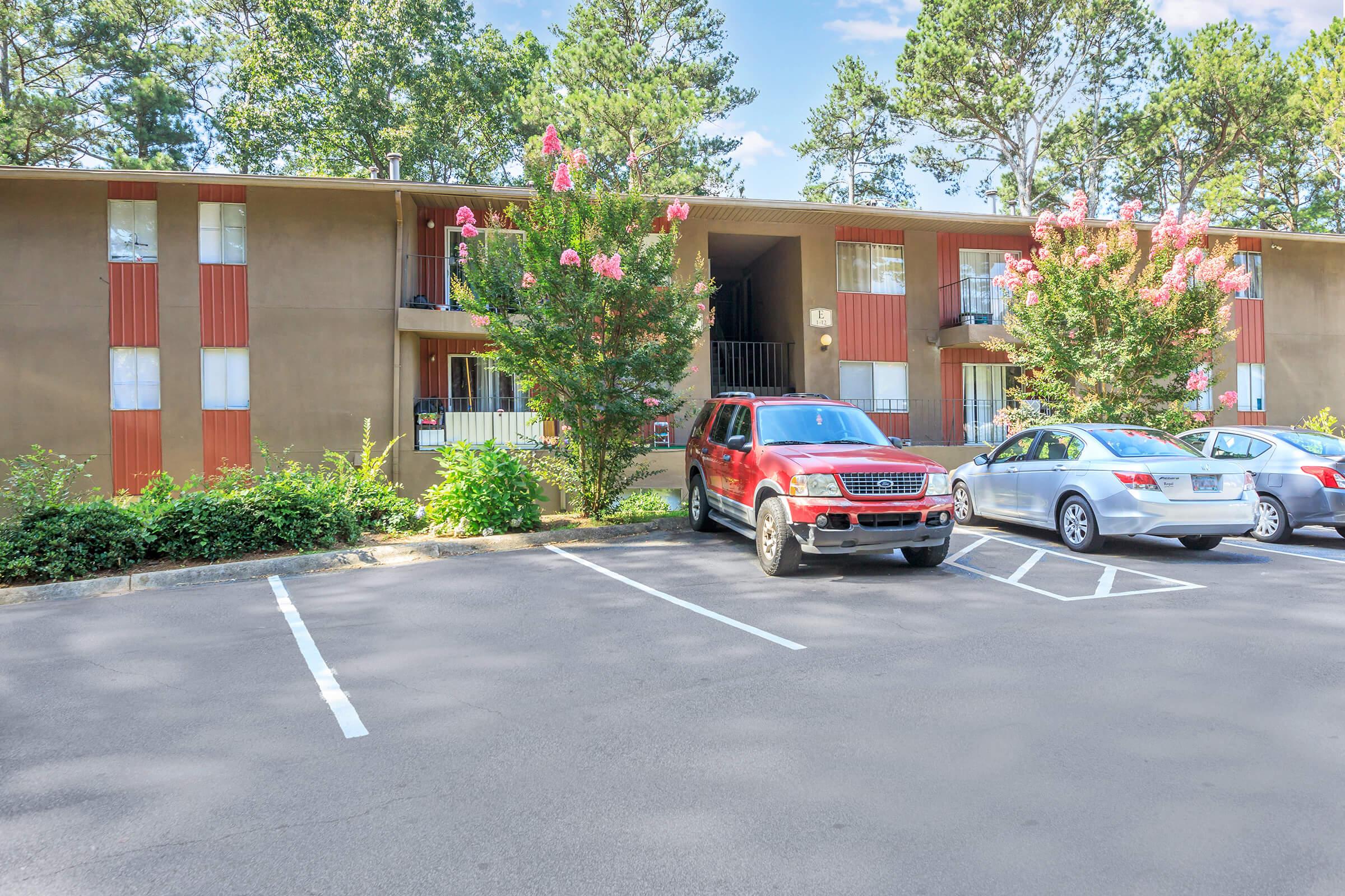 A two-story apartment building with balconies, surrounded by trees. The building features a combination of tan and red exterior colors. In the foreground, there are several parked cars, including a red truck and a silver sedan, with a landscaped area of greenery visible. The atmosphere is sunny and inviting.