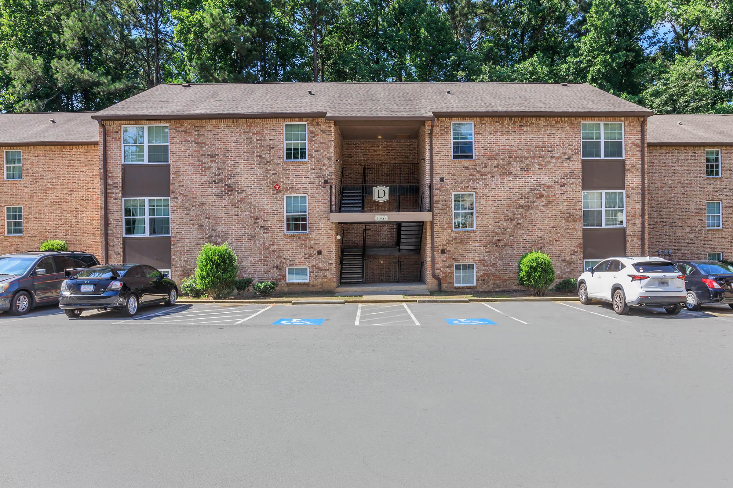 Exterior view of a brick apartment building with multiple units. The building features a central staircase leading to the second floor. Parking spaces, including designated handicapped spots, are in front of the building. Lush greenery surrounds the entrance, contributing to a welcoming atmosphere.