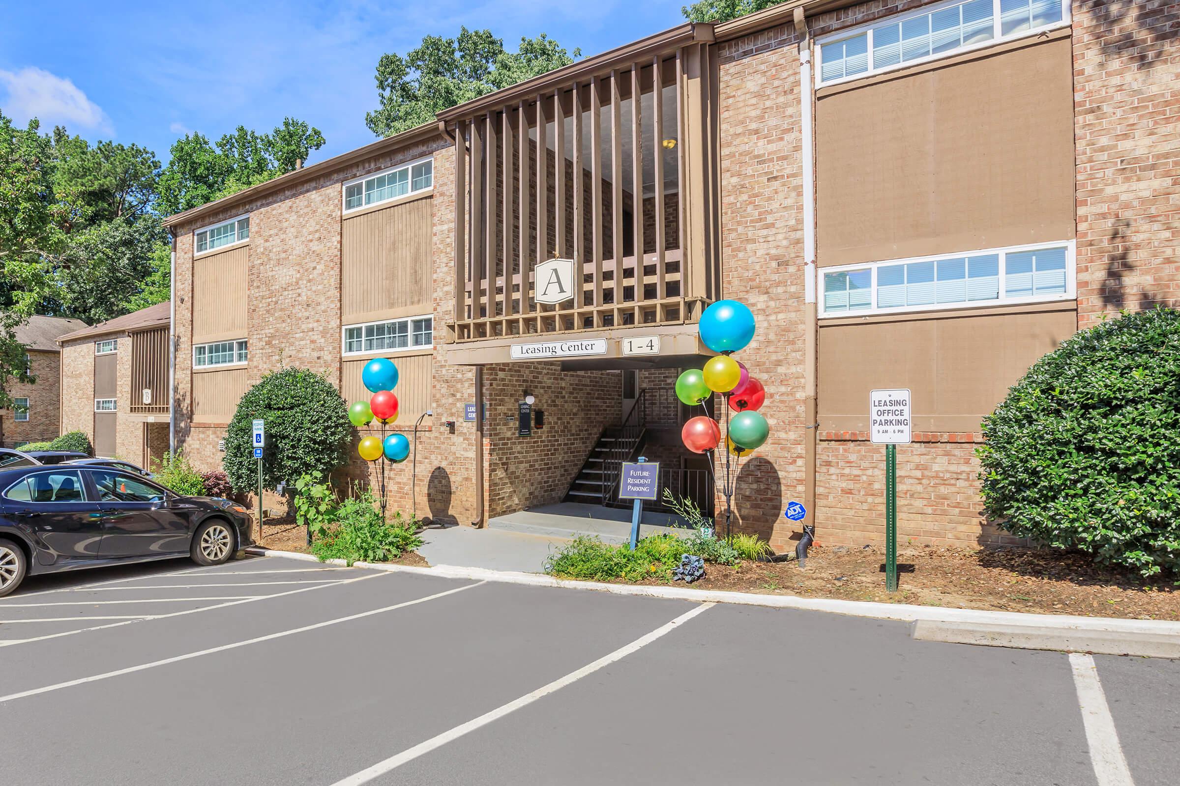 A leasing center entrance of a multi-unit building, featuring balloons in various colors at the entrance. The building has a light brick facade with large windows and a covered area above the entrance. Parking spaces are visible in the foreground.