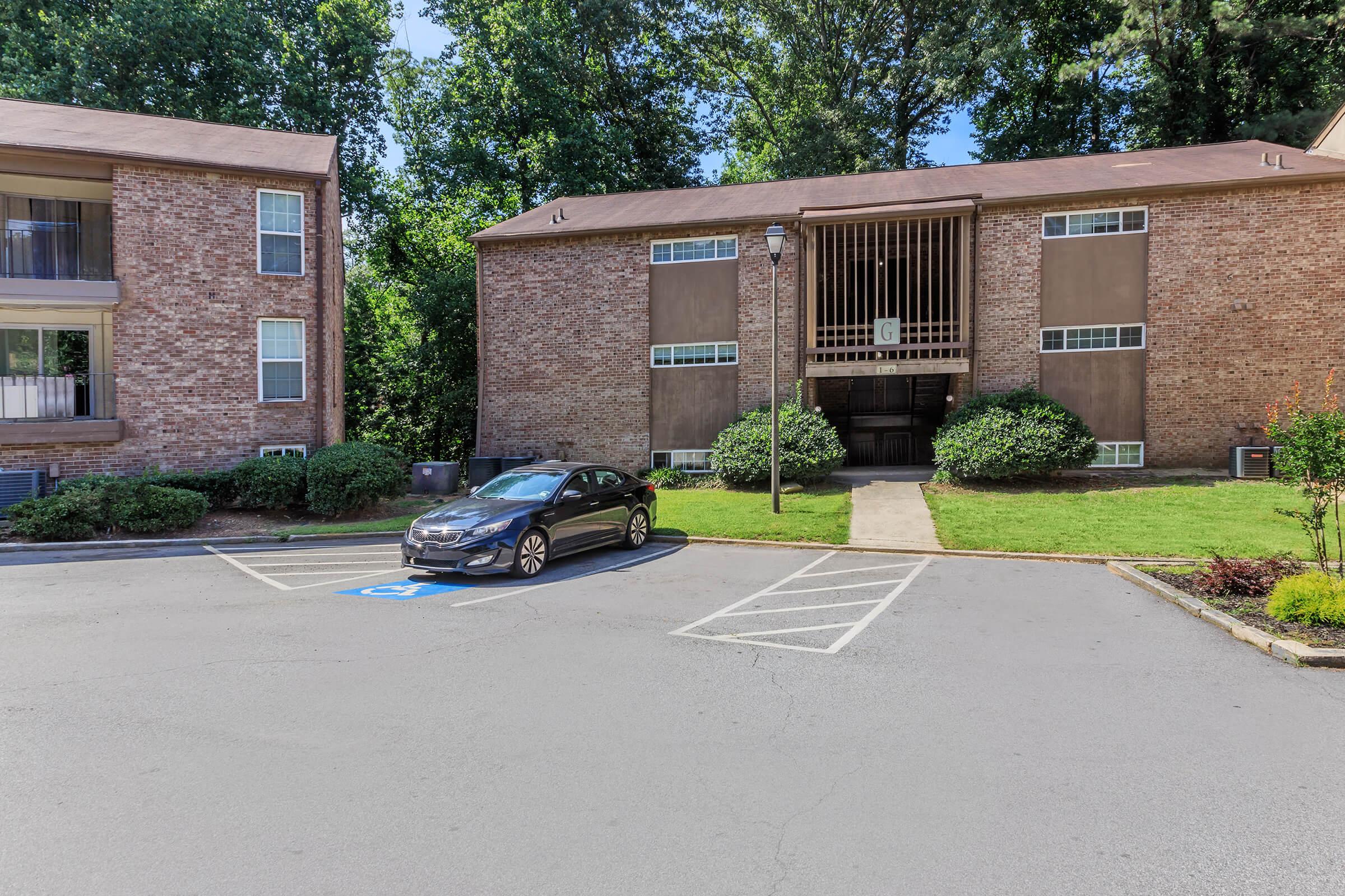 Two brick apartment buildings are shown with a paved parking area in front. A black car is parked in a designated handicapped parking space. The landscape is green with shrubs, and trees are visible in the background. The entrance to one of the buildings is visible, featuring a simple design.