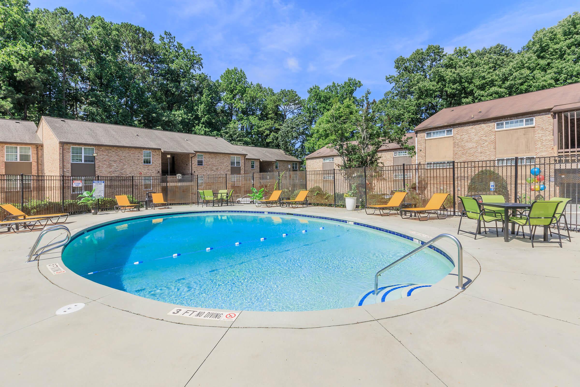 Swimming pool surrounded by lounge chairs and tables, with a fence and trees in the background. Clear blue water reflects a bright sky. The area is well-maintained and inviting, suitable for relaxation and outdoor gatherings.