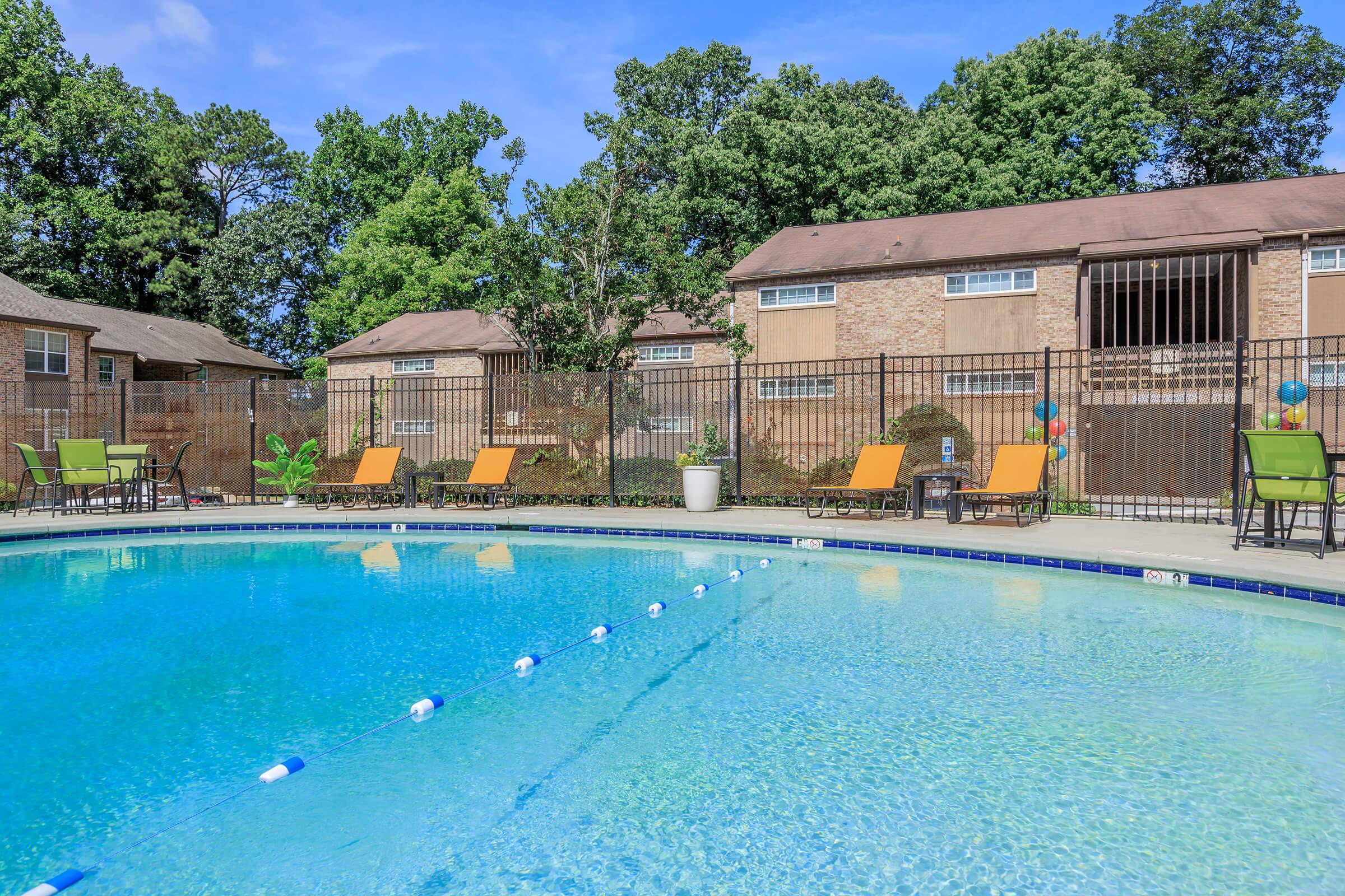 A sparkling blue swimming pool surrounded by green trees and a fenced area. Light brown lounge chairs are positioned around the pool, and colorful decorations add a festive touch. In the background, there are brick apartment buildings with large windows. The scene is bright and inviting, perfect for relaxation.