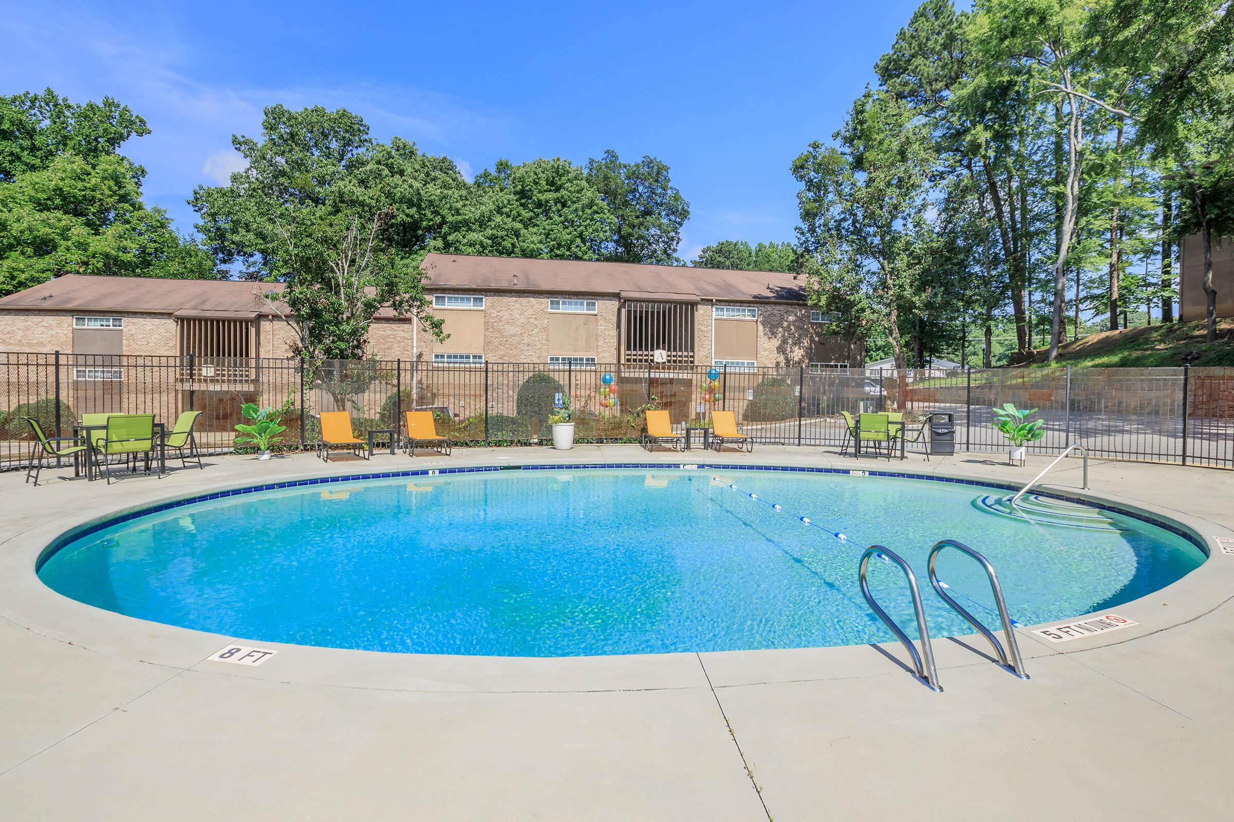 A sunny outdoor swimming pool surrounded by trees, featuring lounge chairs and a secure fence. The pool has a clear blue water and a depth marker indicating 8 feet. In the background, there are brown apartment buildings partially visible.