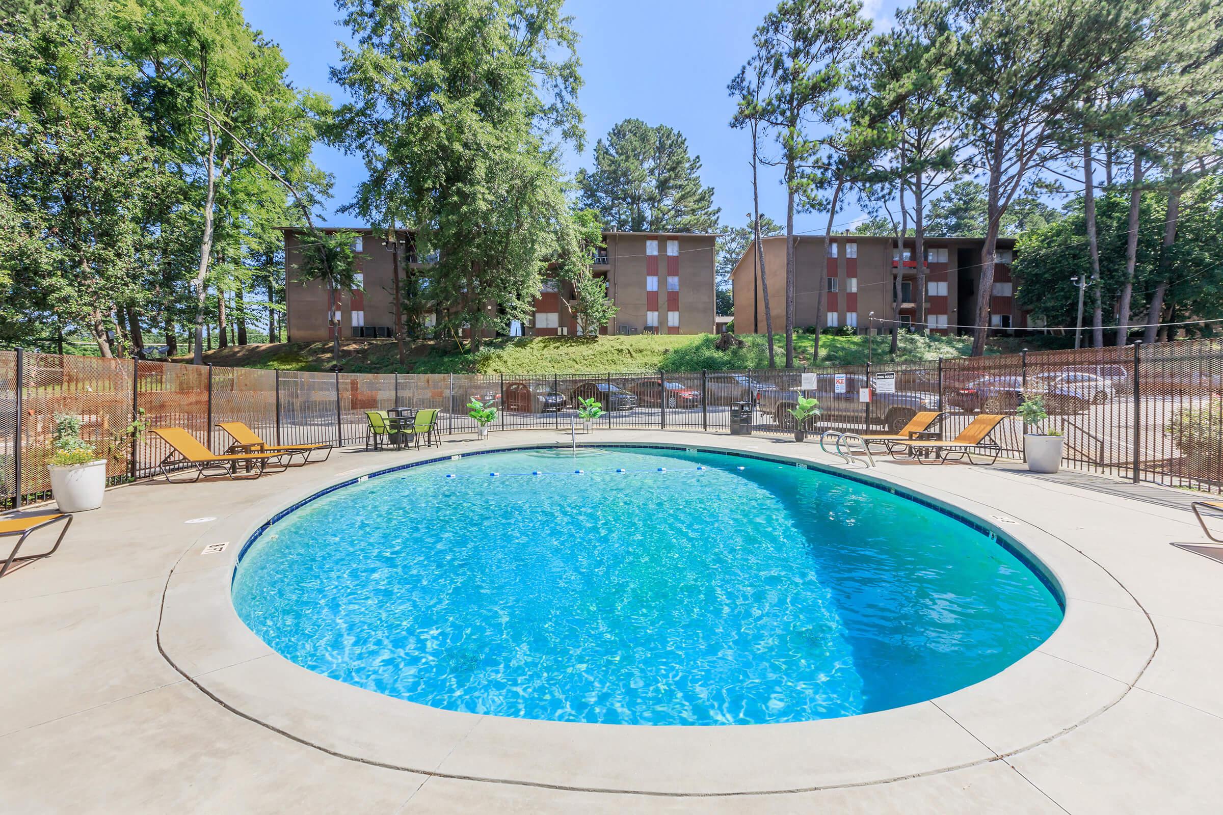 A clear blue swimming pool surrounded by lounge chairs, set in a fenced area with green trees in the background. In the distance, several apartment buildings are visible. The sky is bright blue and sunny, creating a welcoming outdoor environment.