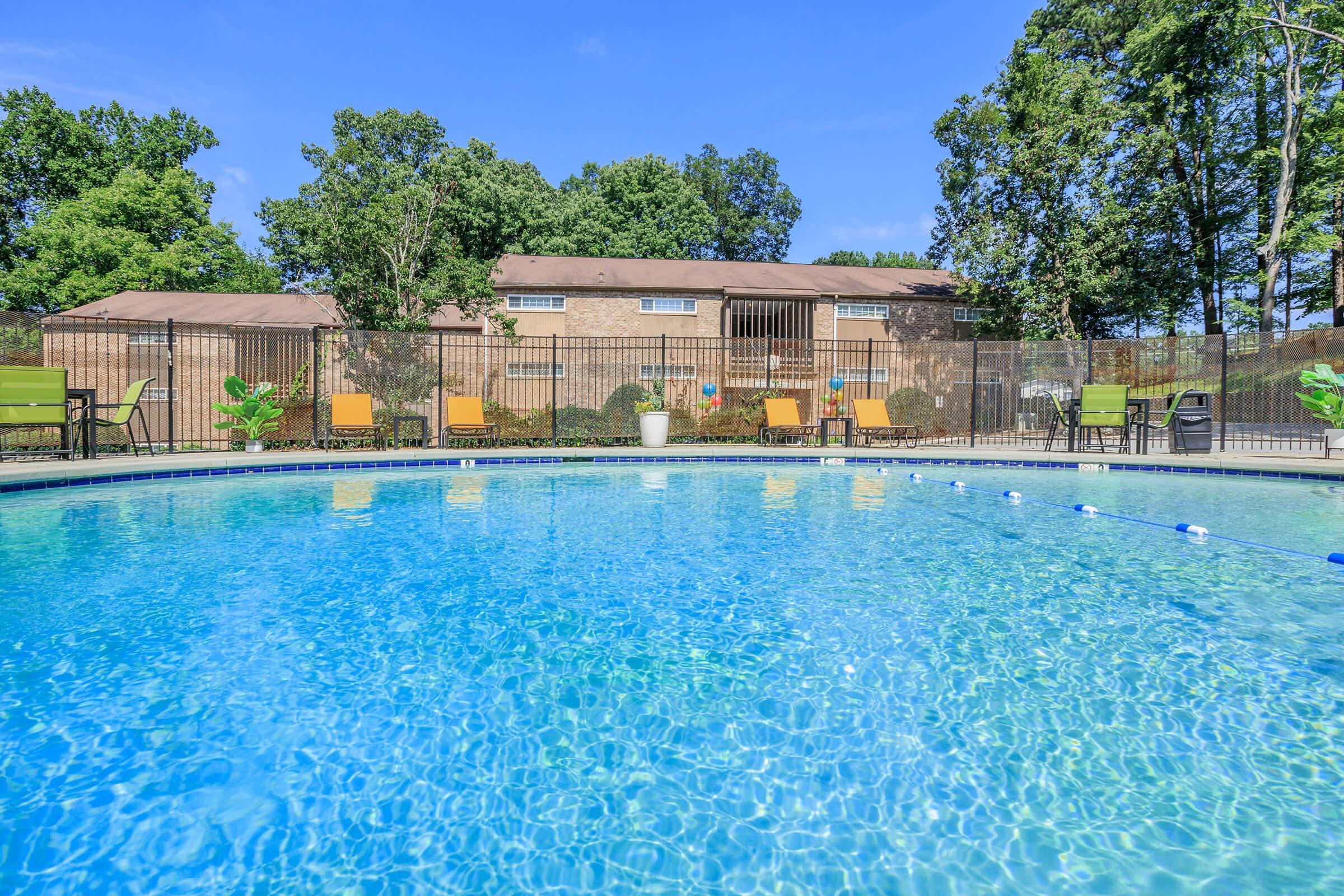 A clear blue swimming pool surrounded by green trees and a wooden fence. Lounge chairs in yellow are positioned around the pool, offering a relaxing space. In the background, there are brown buildings with multiple windows, set against a bright blue sky.
