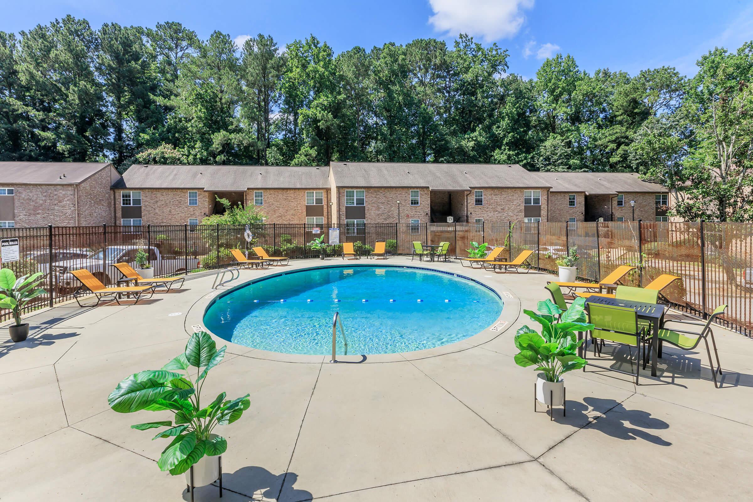 A vibrant outdoor pool area featuring a circular swimming pool surrounded by yellow lounge chairs, green plants, and tables. In the background, there are brick apartment buildings framed by lush greenery under a bright blue sky.