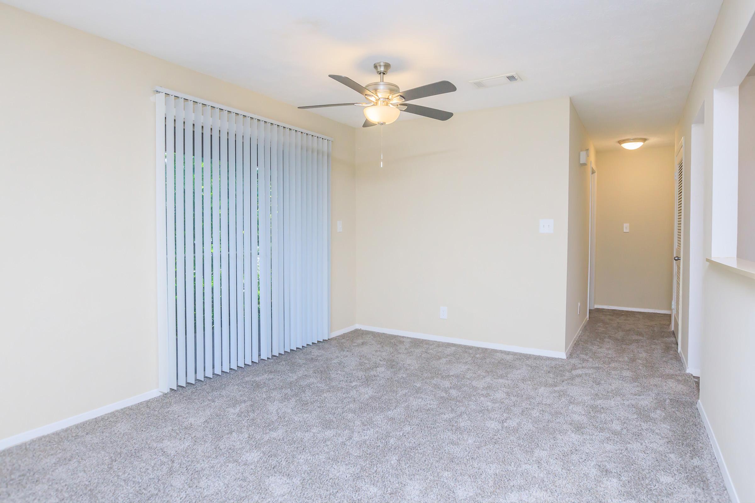 A spacious, light-filled living room with beige walls and plush gray carpet. It features a ceiling fan and vertical blinds on a sliding glass door that offers access to an outdoor area. The room has a neutral color scheme and a doorway leading to a hallway.