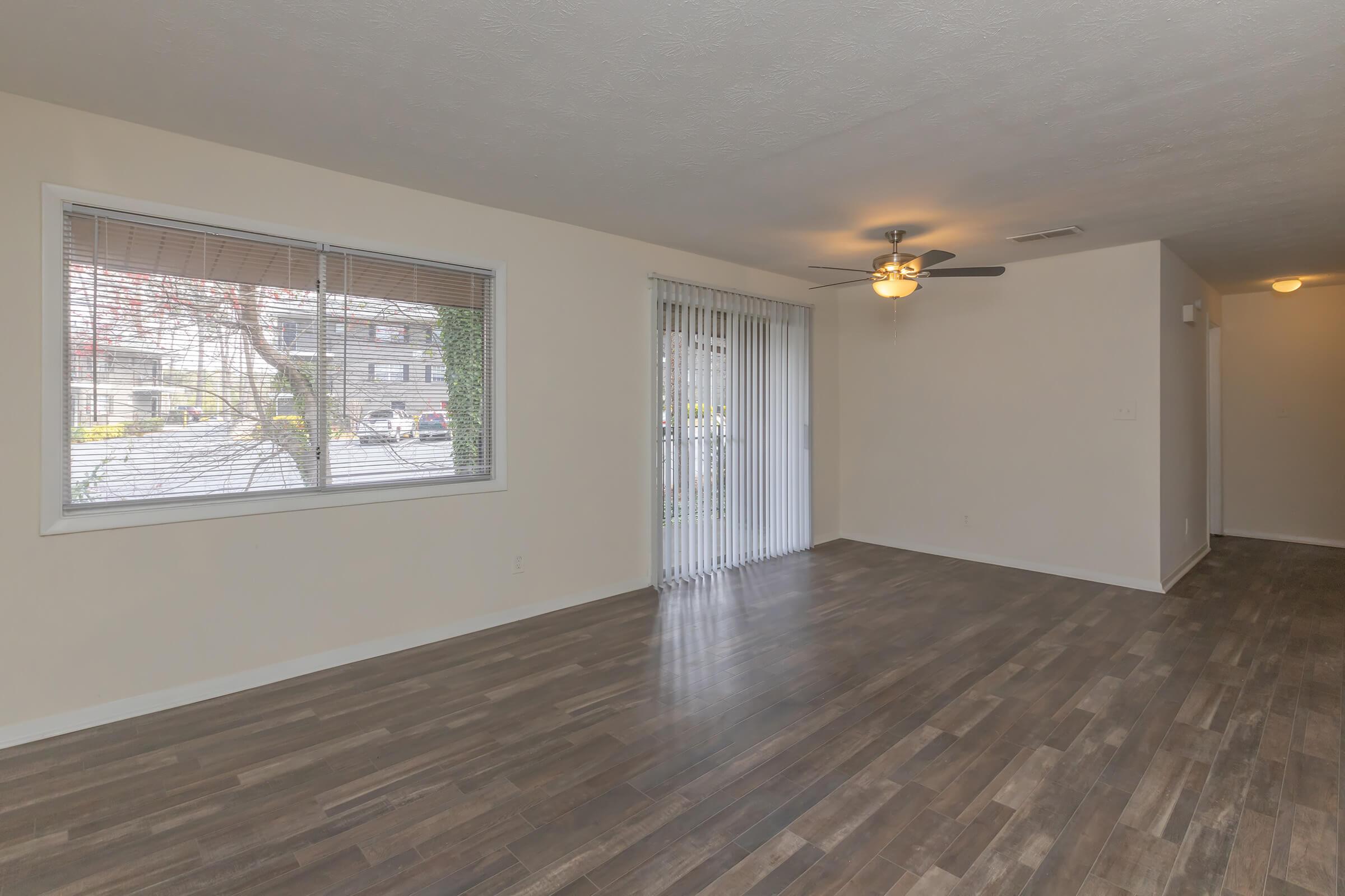 A spacious, empty living room with hardwood-like flooring, a ceiling fan, and large windows allowing natural light. There are vertical blinds on the sliding door, leading to an outdoor area, and neutral-colored walls enhance the sense of openness.