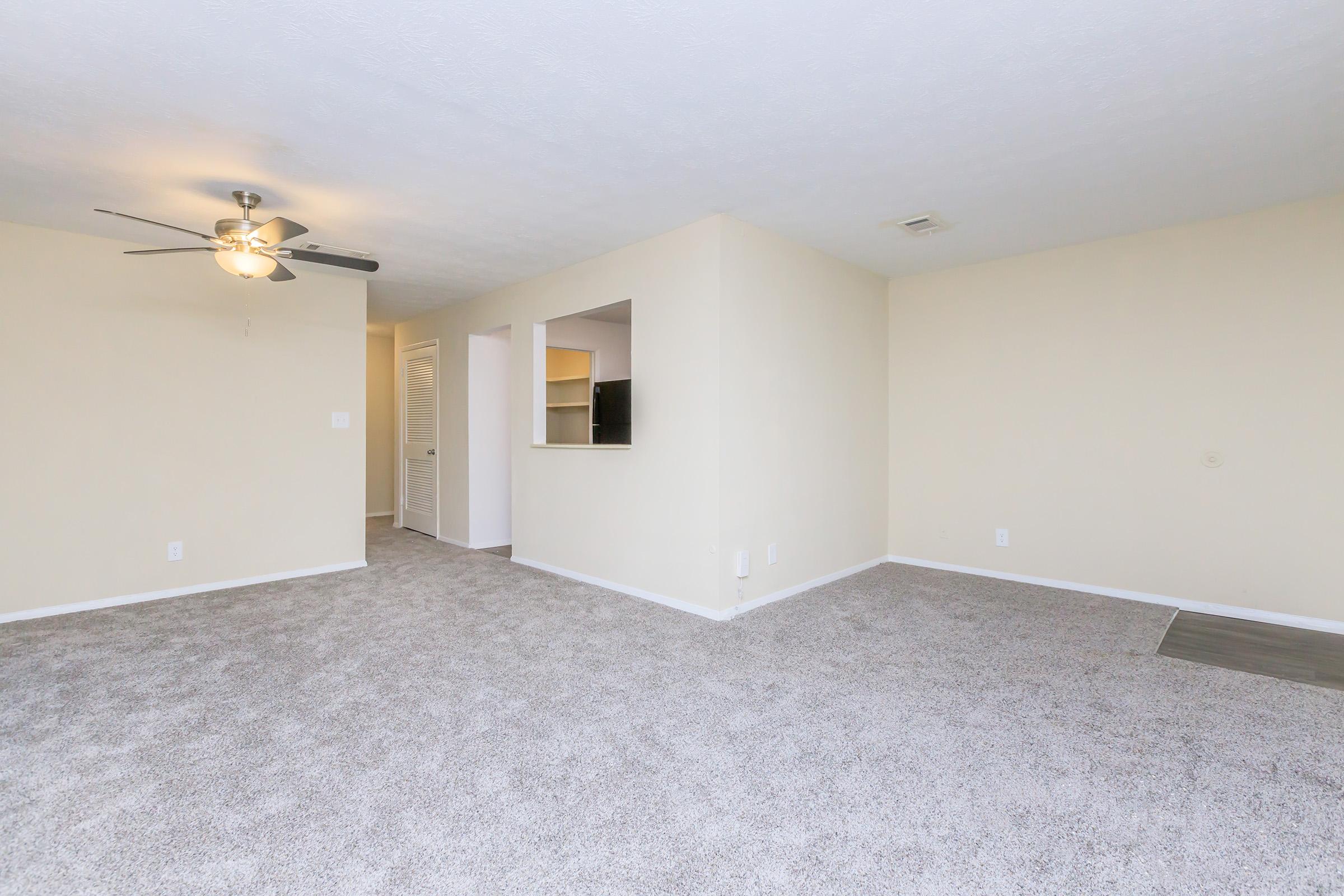 Spacious, light-filled living area with beige walls and plush carpet. Features a ceiling fan and an open layout leading to a small kitchen area visible through an archway. A doorway to a hallway is seen on the left, creating a welcoming atmosphere for relaxation or entertaining. Ideal for comfortable living.