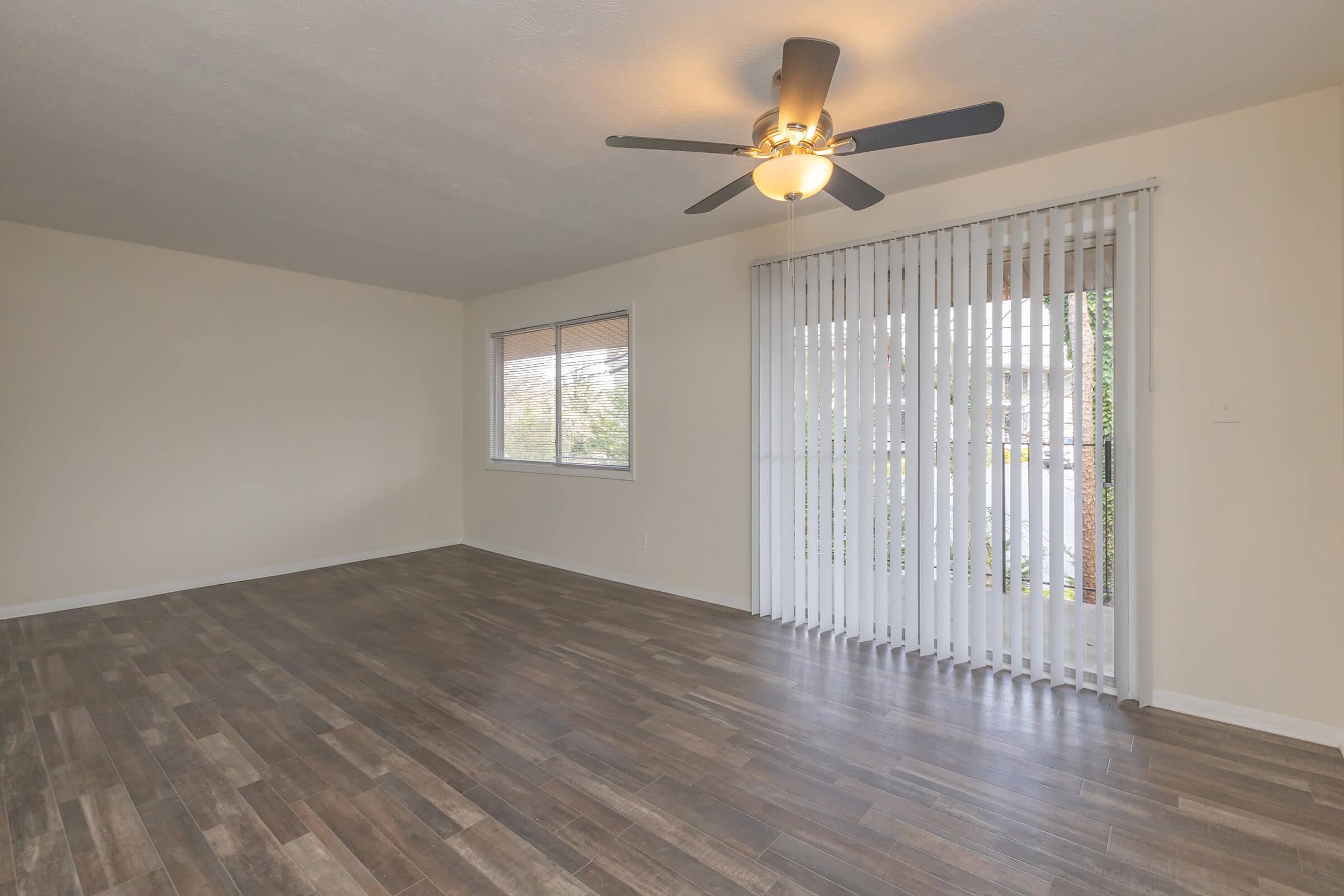 Spacious, empty living room with light-colored walls and a ceiling fan. Features large windows with vertical blinds, allowing natural light. The floor is covered in dark wood laminate, creating a warm ambiance. A sliding glass door opens to an outdoor area, enhancing the room's airiness.