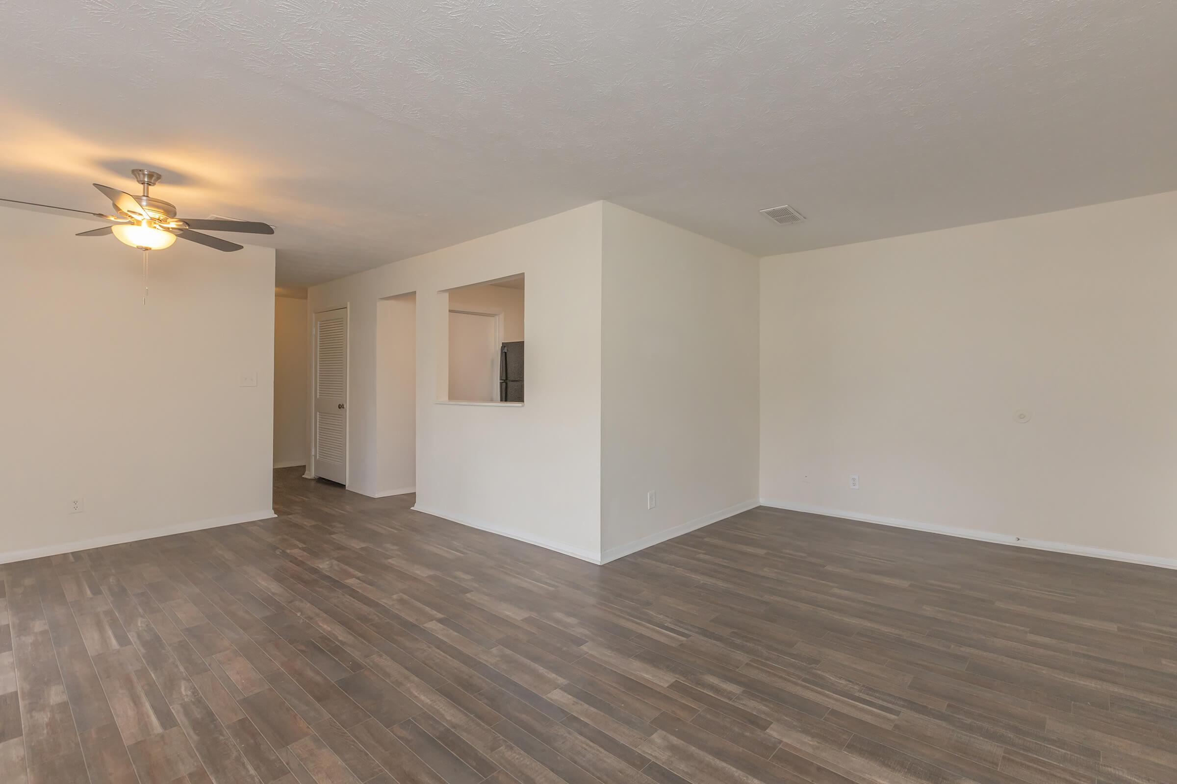 A spacious, empty living area with light-colored walls and wood-style flooring. A ceiling fan is present, and there is an opening leading to a hallway and kitchen area. Natural light illuminates the room, creating a bright atmosphere.