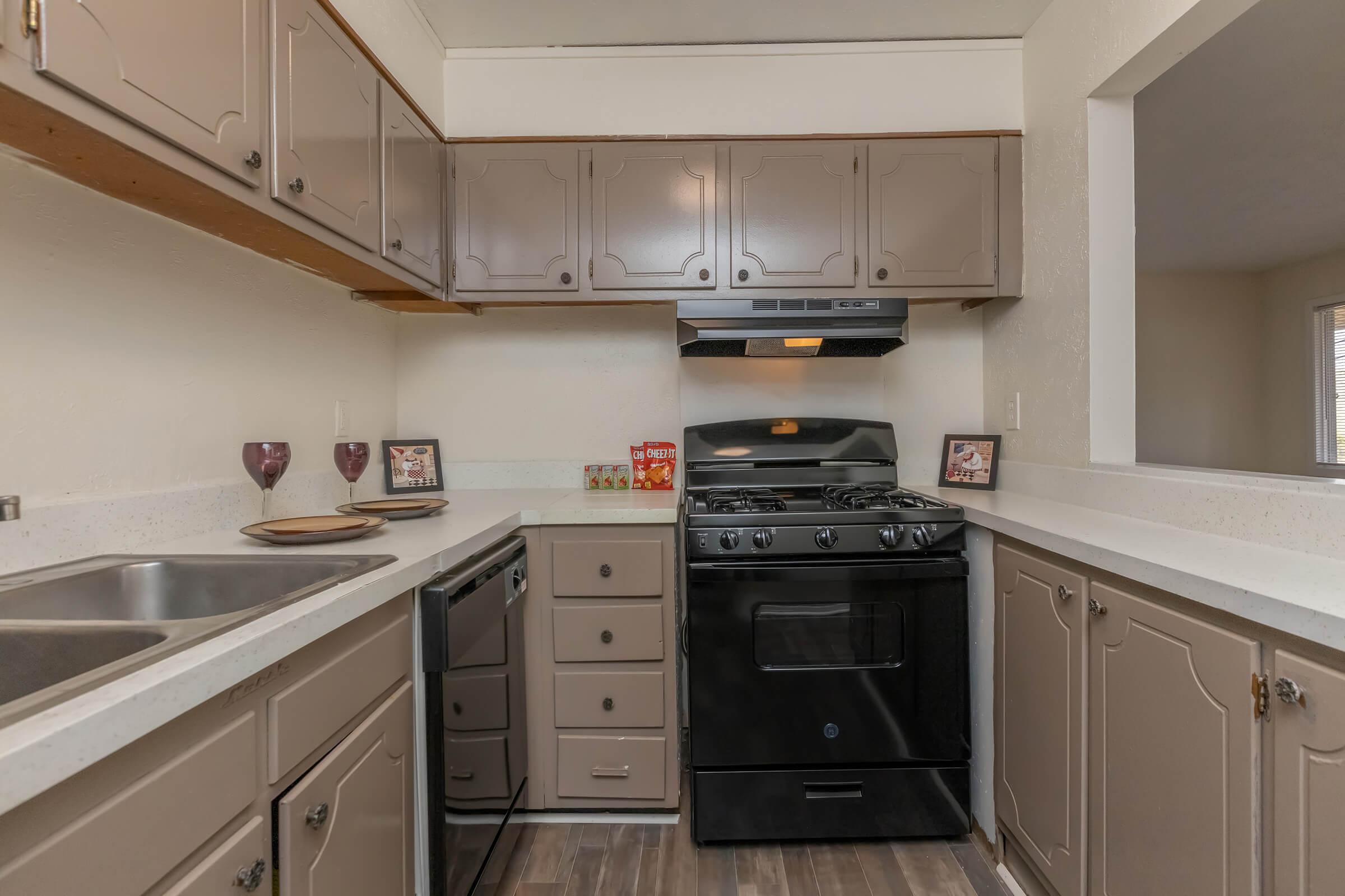 A modern kitchen featuring beige cabinets, a black stove, and a black dishwasher. The countertop is light-colored, and there are decorative items and wine glasses on the counter. The layout is compact, with a window offering natural light.