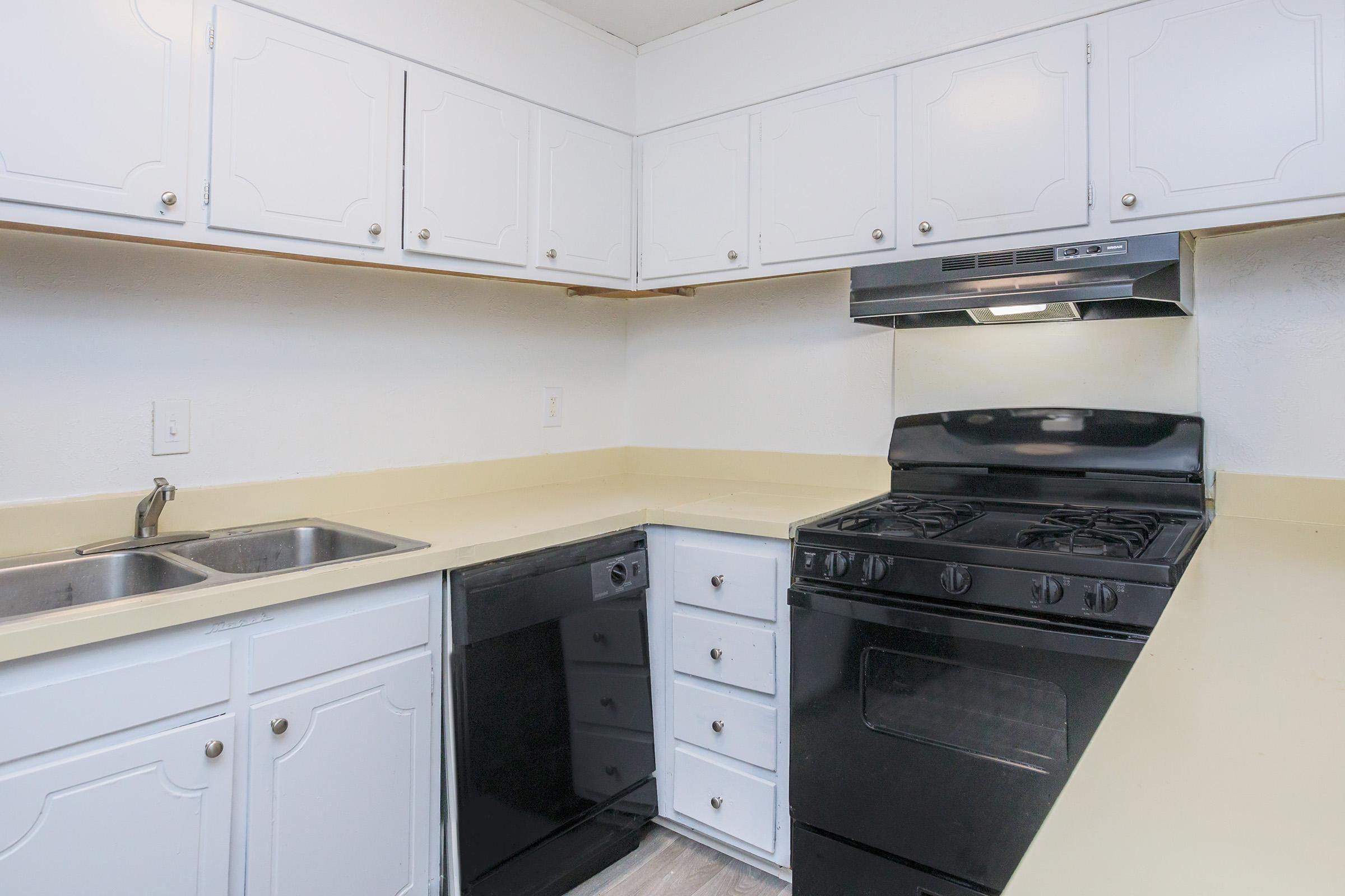 A clean, modern kitchen featuring white cabinetry, a beige countertop, a black stove, and a black dishwasher. Two sinks are installed under a window. The overall design is simple and functional, with a bright, uncluttered appearance.