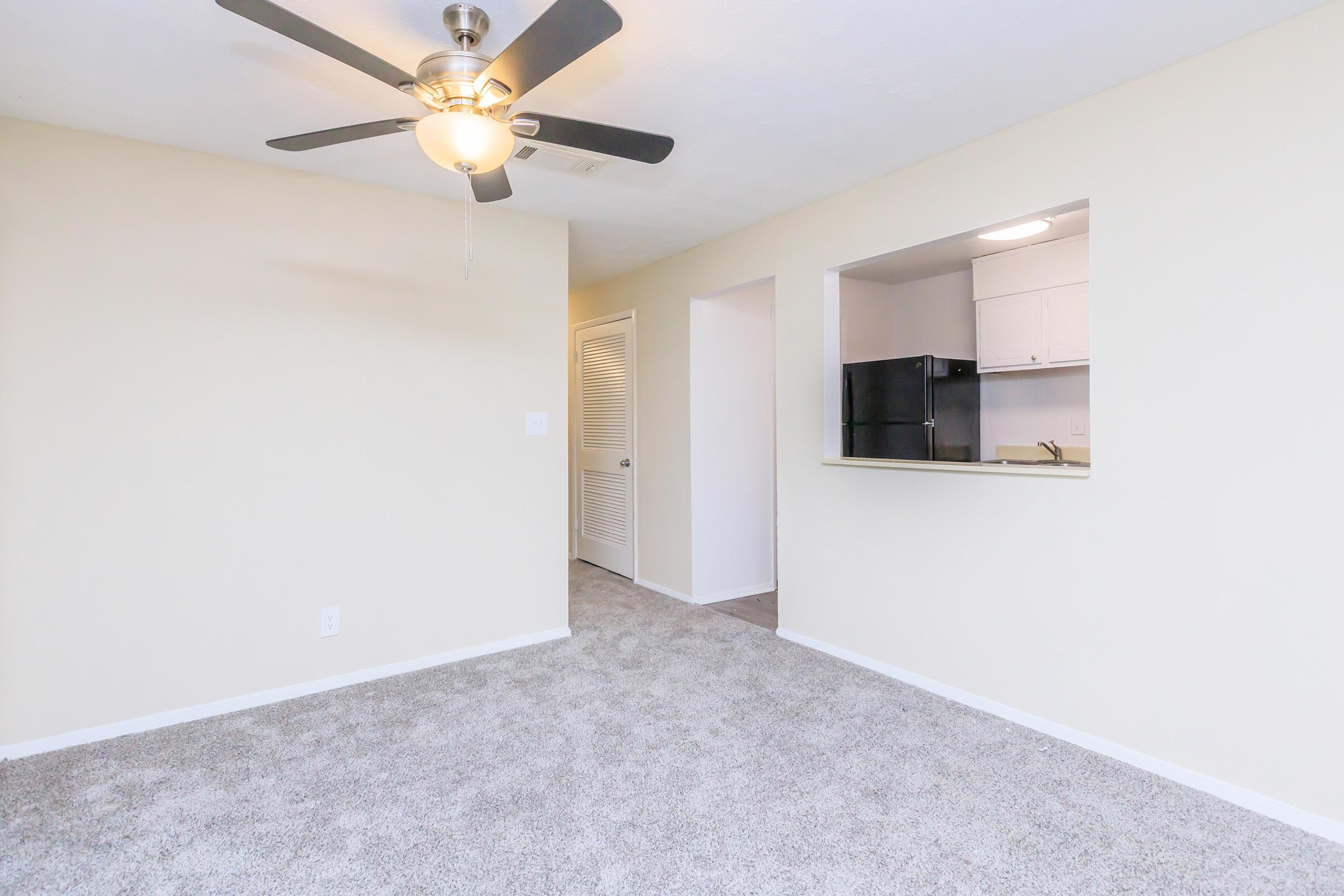 A spacious living area with light beige walls and carpet, featuring a ceiling fan. An open kitchen is visible in the background, with white cabinetry and a black refrigerator. A door leads to another room, enhancing the open layout. Natural light fills the space, creating a welcoming atmosphere.
