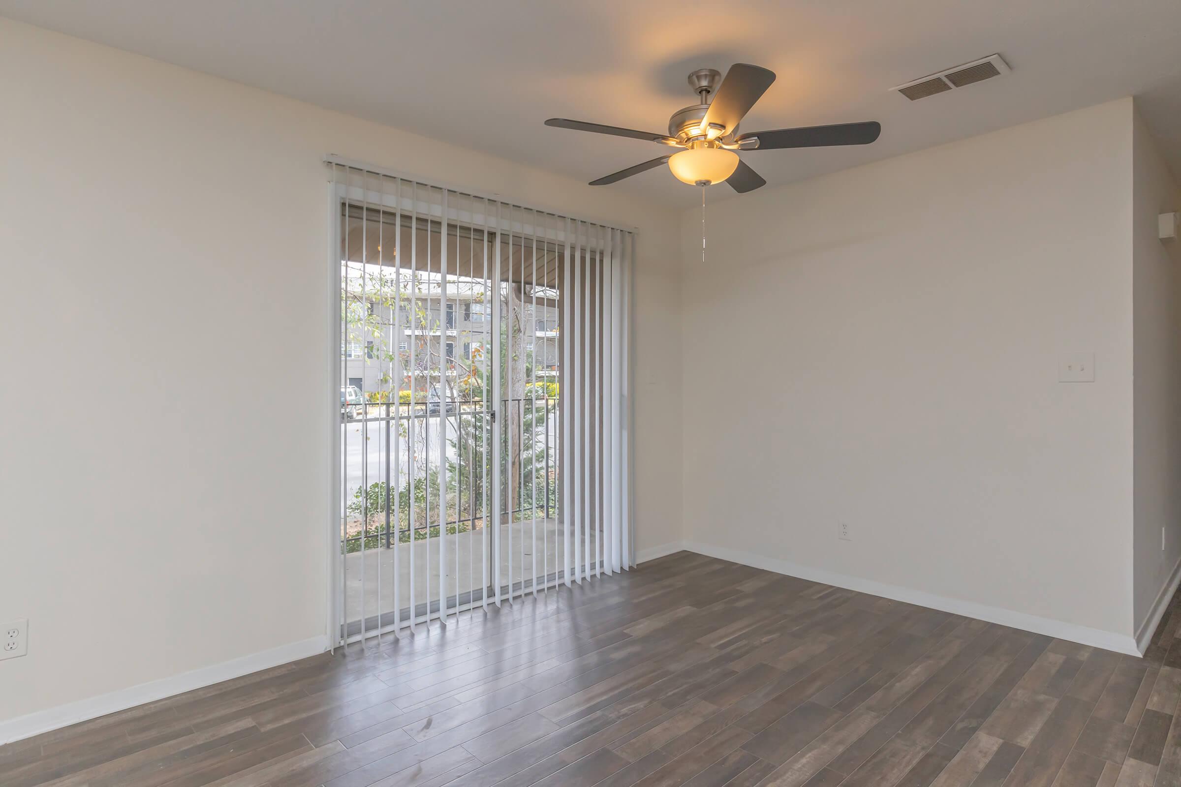 A spacious, empty living room with brown wooden floors, white walls, and a ceiling fan. Large sliding glass doors with vertical blinds lead to an outdoor area. Natural light fills the room, creating a bright and inviting atmosphere, ideal for decorating or personalizing with furnishings.