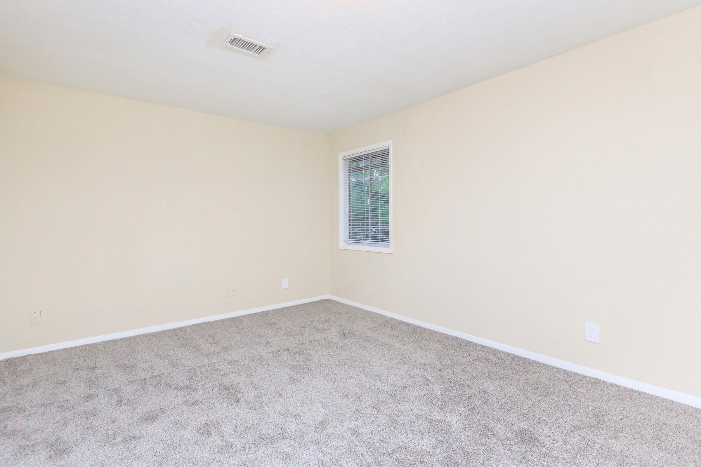 Empty room with beige walls and neutral-colored carpet. A single window allows natural light to enter, and there are no furnishings or decorations present. The space appears clean and well-maintained, suitable for various uses.