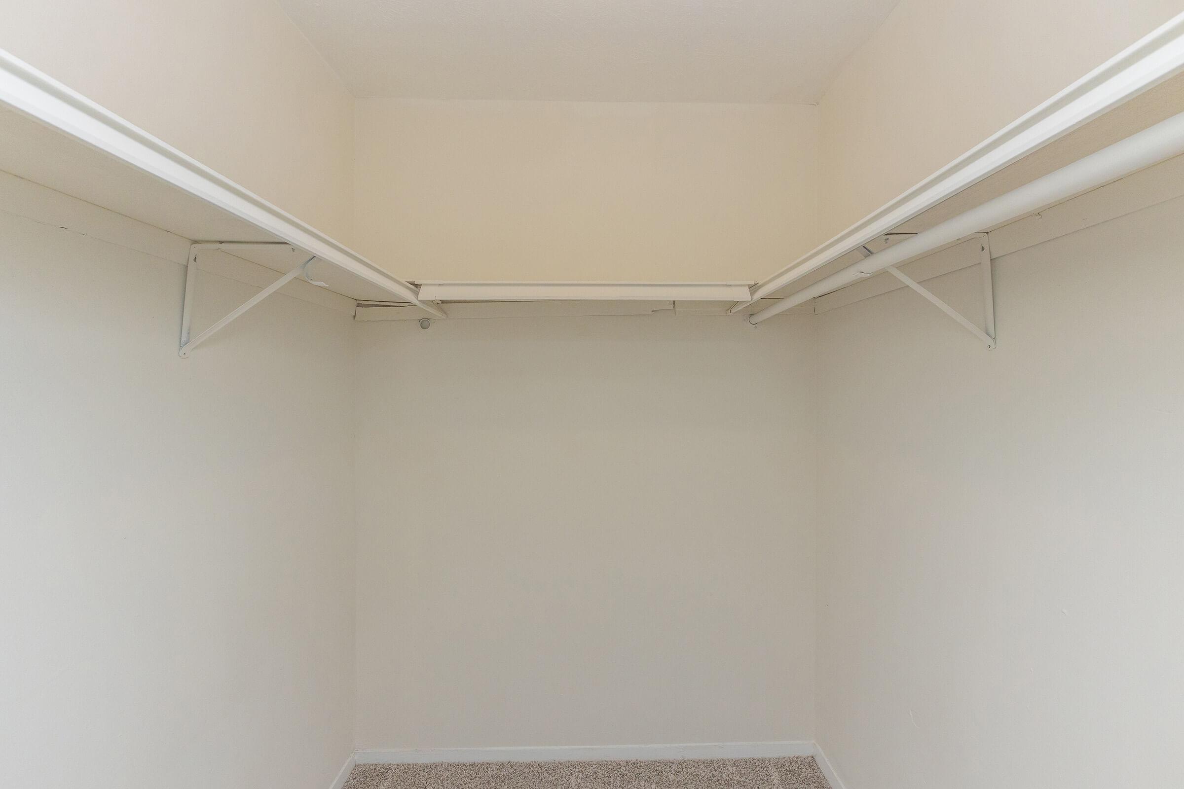 Empty closet with beige walls and a light carpet. The closet features two horizontal hanging rods supported by metal brackets, providing storage space for clothes and accessories. The overall design is minimalistic and unadorned.