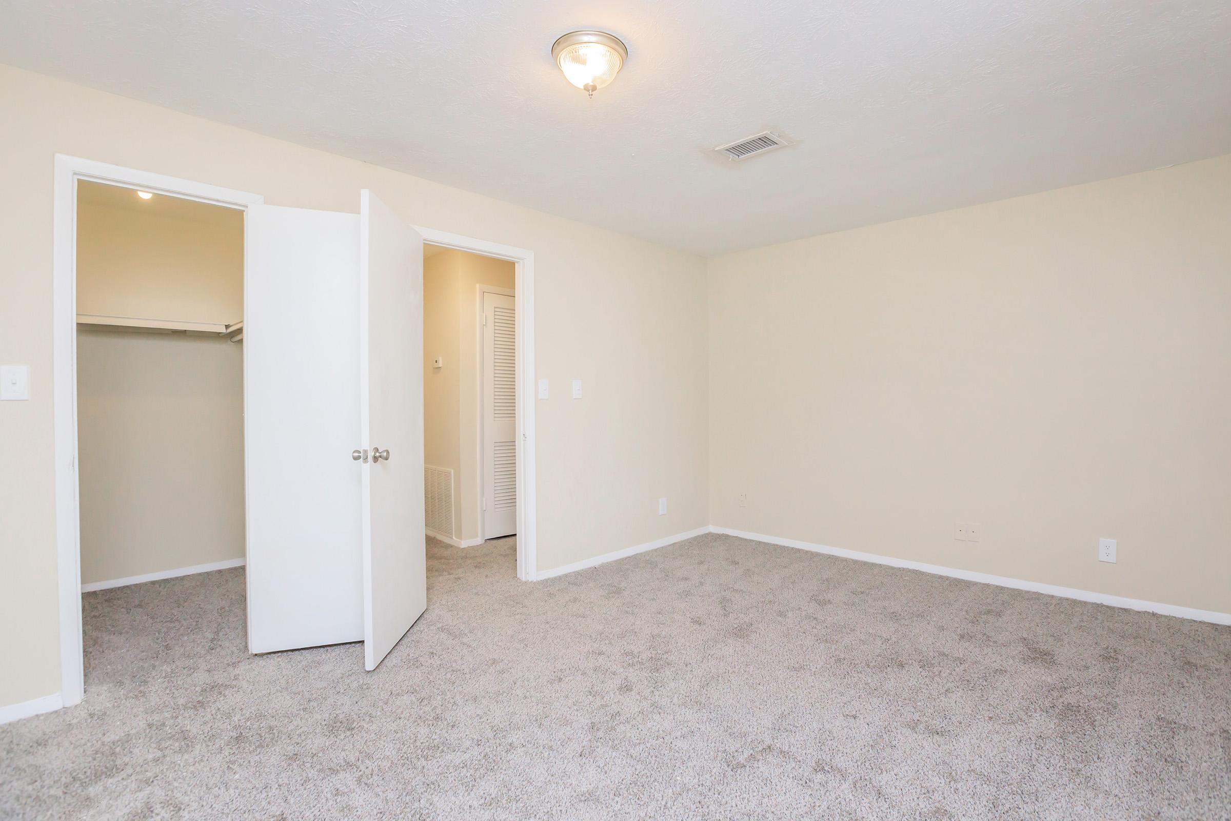 A spacious, empty bedroom featuring light beige walls and plush gray carpeting. One door is ajar, leading to a closet, while another door opens to a hallway. The room is well-lit with a ceiling light fixture, providing a bright and inviting atmosphere.
