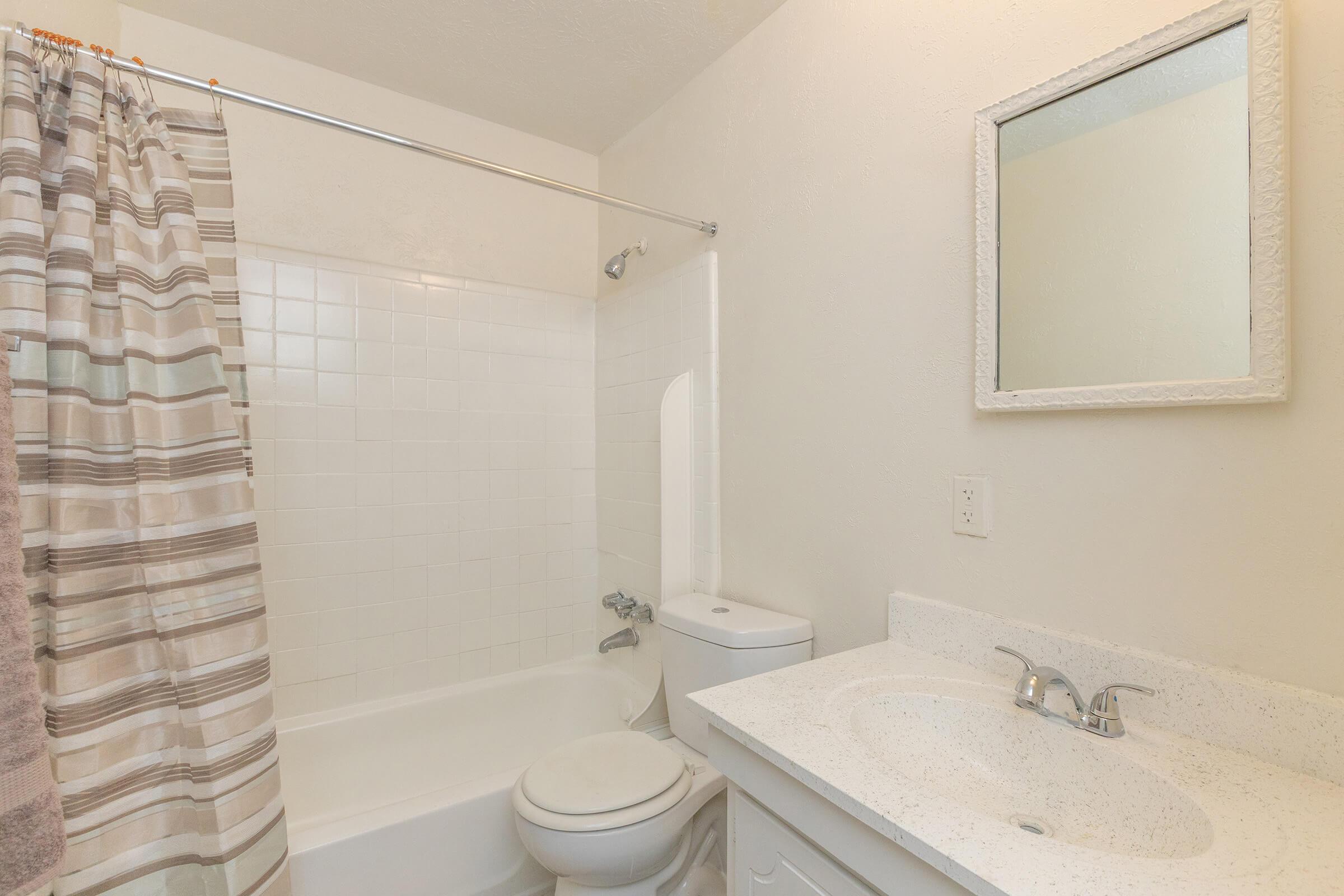 A clean, well-lit bathroom featuring a bathtub with a shower curtain, a toilet, and a sink with a countertop. The walls are painted in a light color, and a small framed mirror hangs above the sink. The shower curtain has a striped design, and fluffy towels are visible hanging nearby.