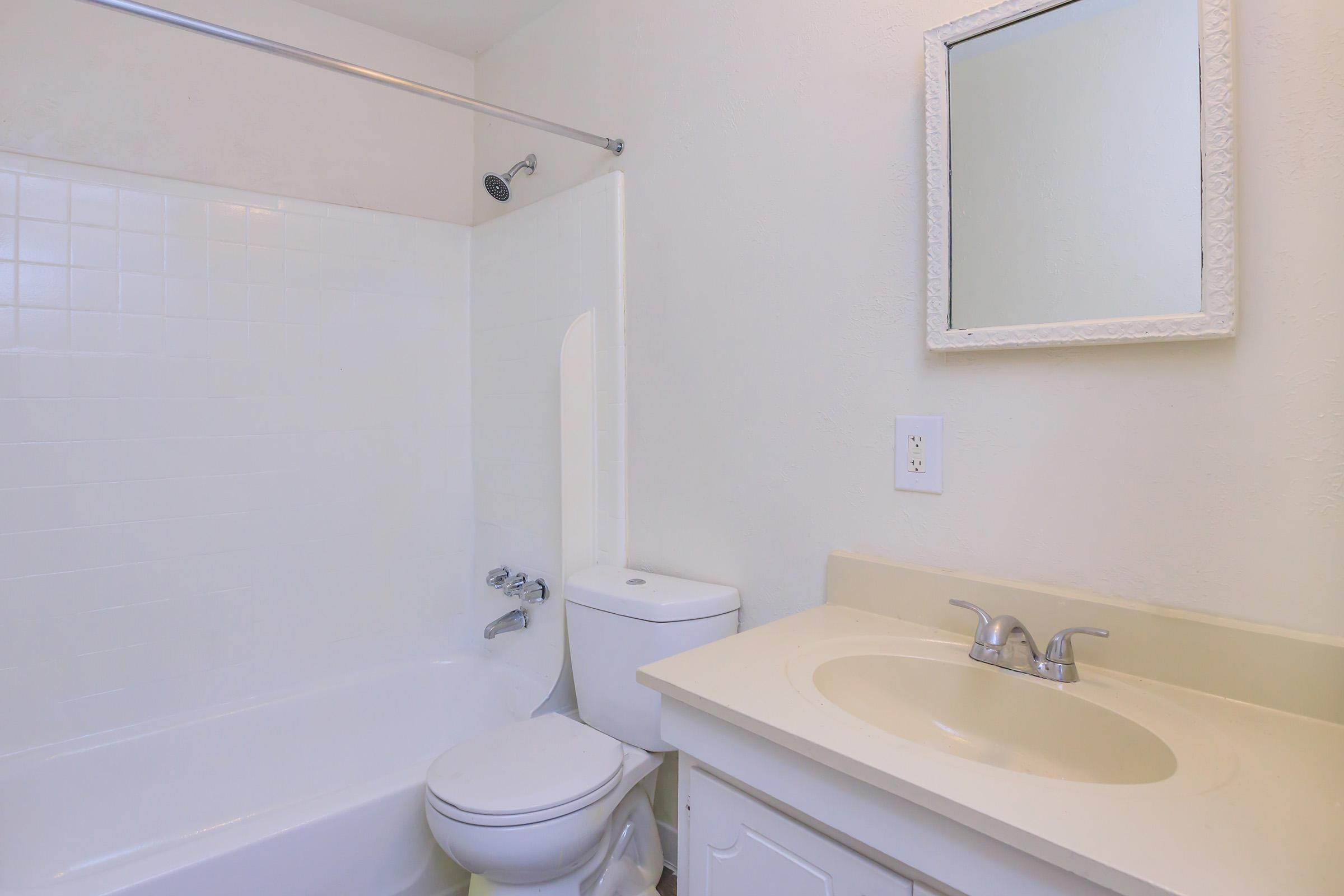 A clean, bright bathroom featuring a white bathtub, a toilet, and a sink with a light-colored countertop. The walls are painted in a neutral tone, and there is a mirror mounted above the sink. The overall appearance is simple and modern, with a functional layout.