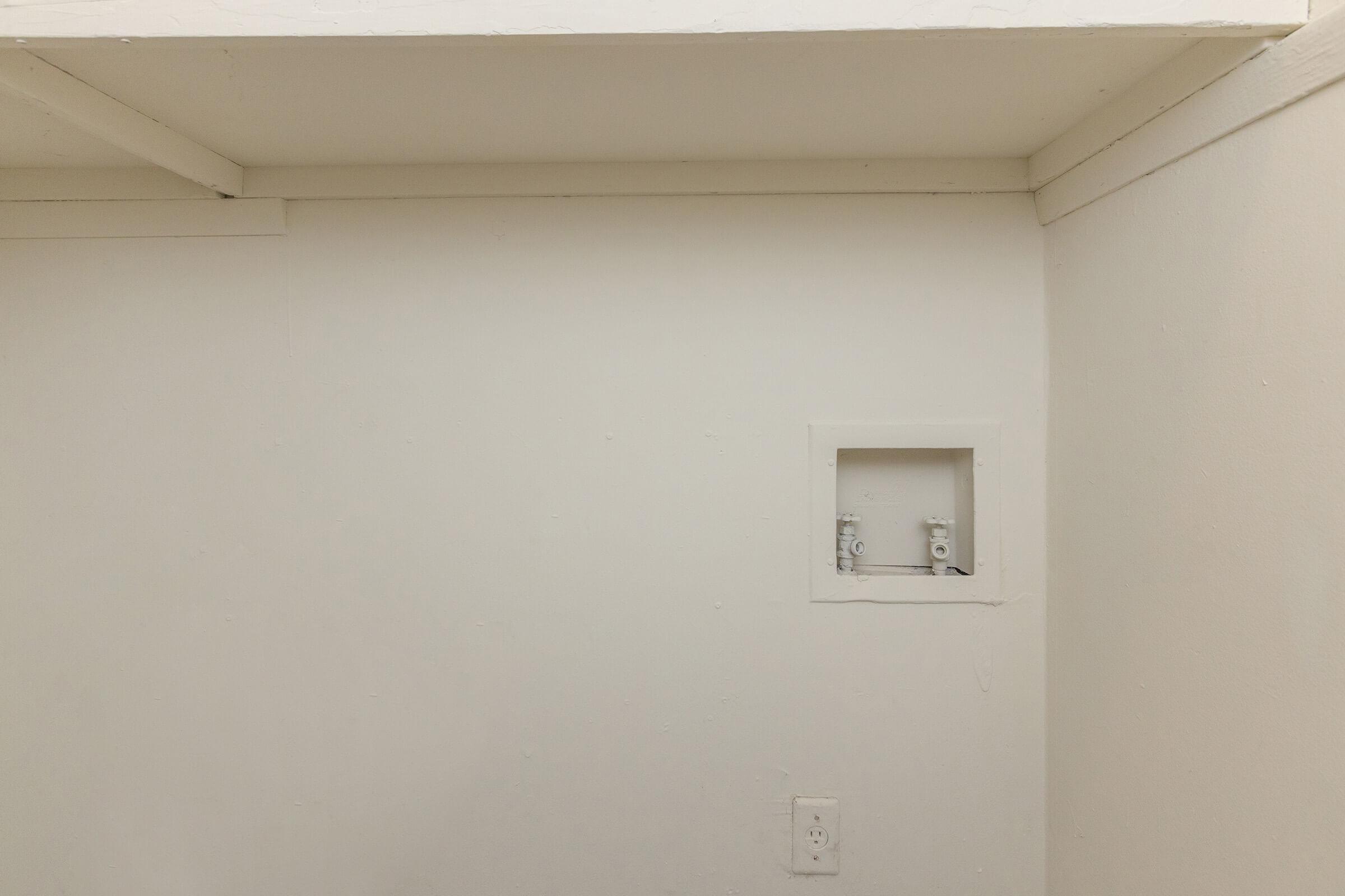 Empty room with white walls and ceiling. A small rectangular opening is visible on the left side, likely for an electrical box. The floor is not visible, and there are no decorations or furniture in the space. Overall, the area appears minimalistic and unfinished.