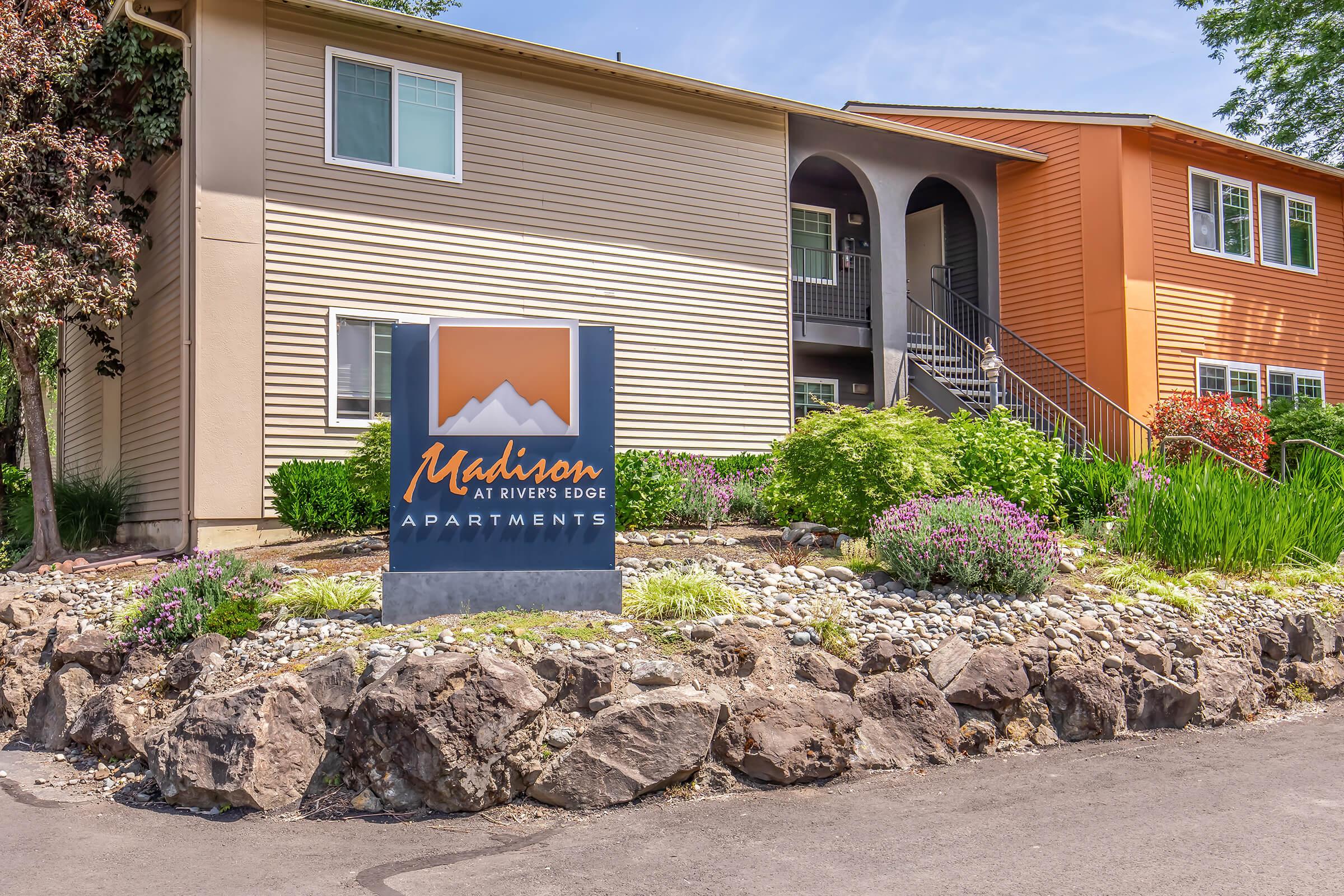 Exterior view of Madison at Rivers Edge Apartments, showcasing the building's entrance and landscaping. A prominent sign with the apartment name is displayed in front, surrounded by rocks and colorful plants. The architecture features different color palettes, highlighting a welcoming atmosphere.
