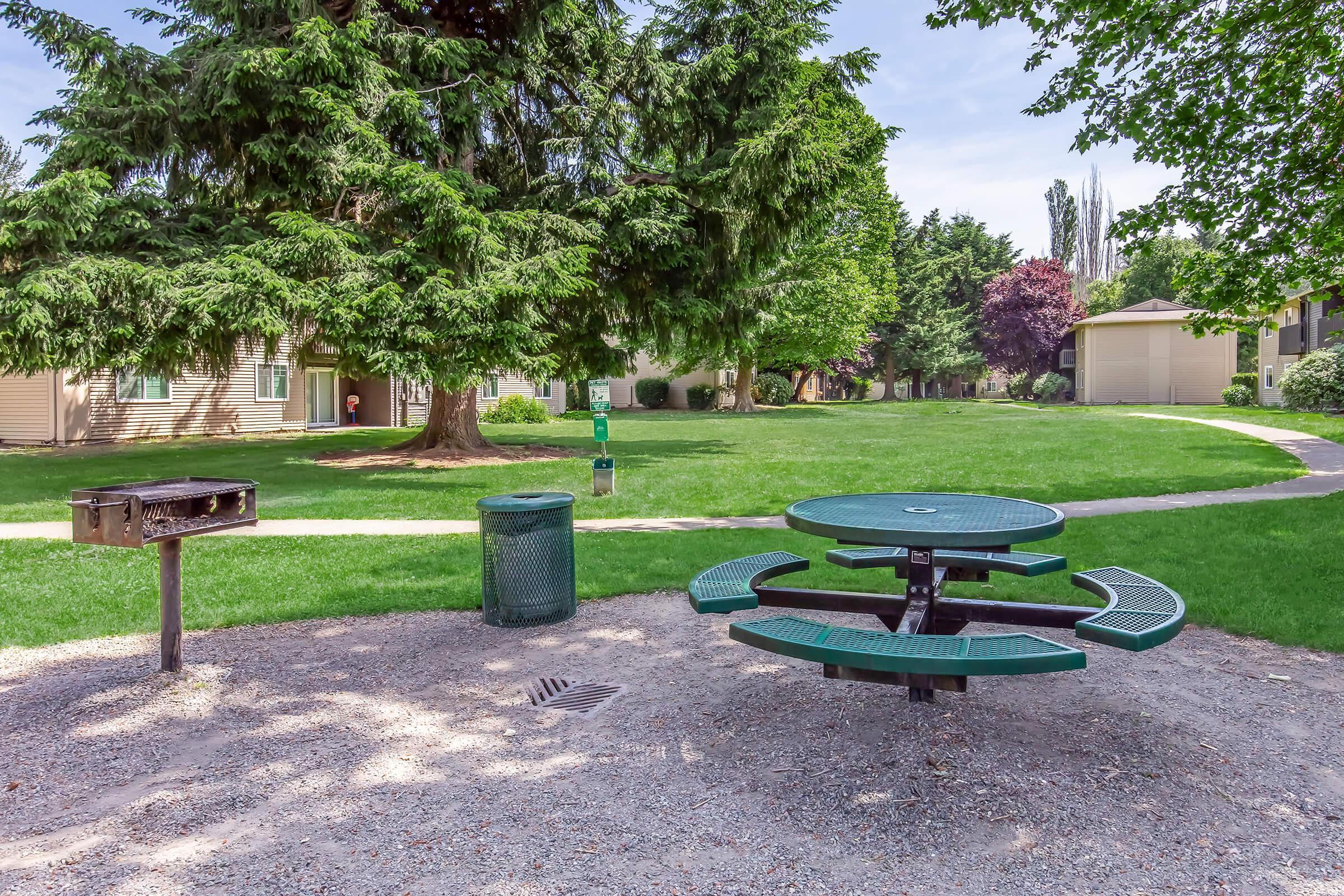 A picnic area featuring a green round table with benches, surrounded by a grassy lawn and trees. A charcoal grill and a trash bin are nearby. Apartment buildings can be seen in the background, and the scene is set on a clear, sunny day.