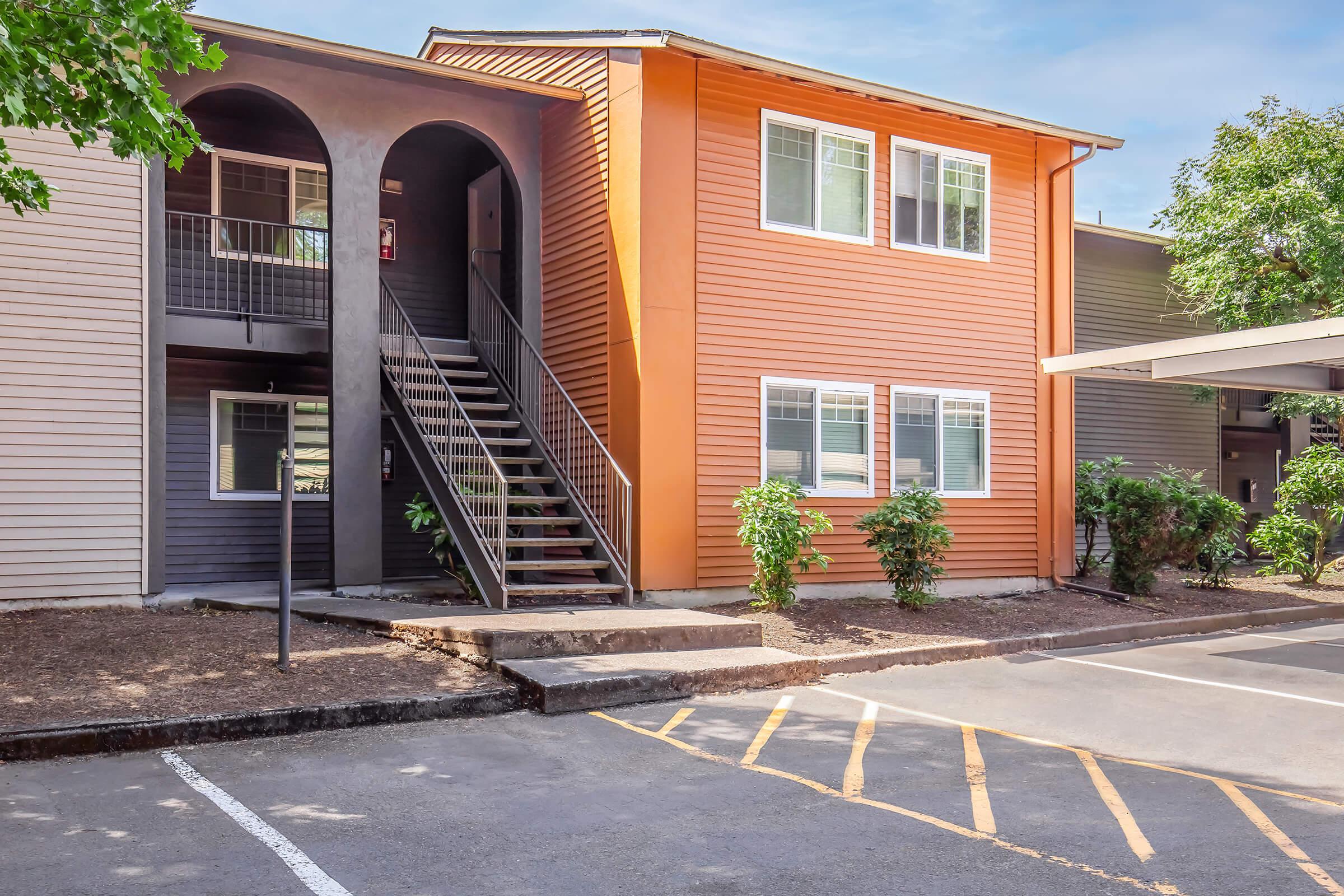 Exterior view of an apartment building featuring a two-tone facade with an orange section. A staircase leads to the upper floor, surrounded by shrubbery. A parking lot is in the foreground, with marked spaces and a covered parking area to the side. The scene is bright, with clear blue skies.
