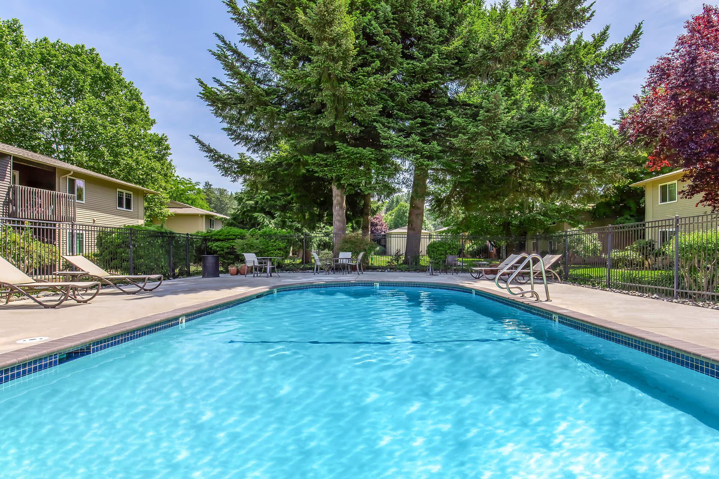 A clear blue swimming pool surrounded by lush green trees and landscaping. Lounge chairs are positioned around the pool area, and there are tables in the background. The scene is set in a residential community, showcasing a peaceful and inviting outdoor space.