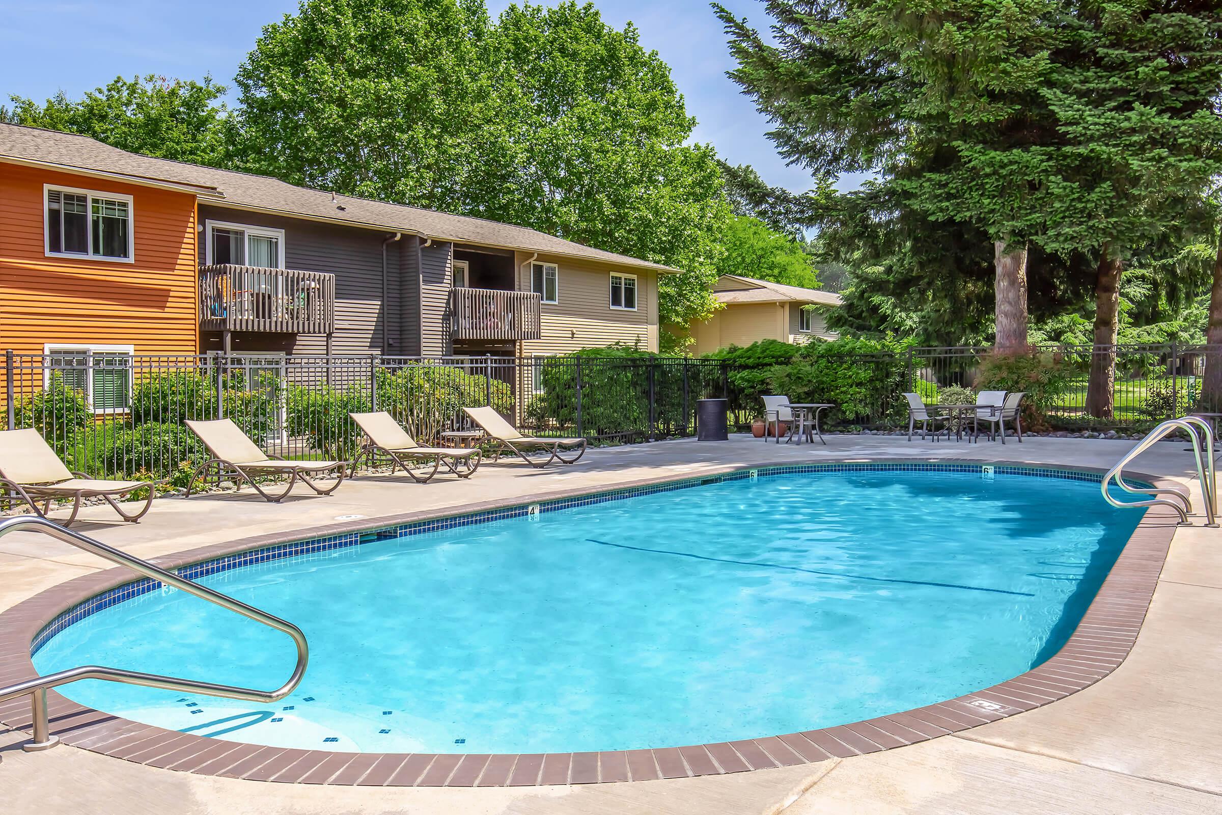 A tranquil outdoor swimming pool surrounded by lounge chairs, with nearby trees and colorful apartment buildings in the background. The clear blue water reflects the sunny sky, creating a relaxing atmosphere perfect for leisure and enjoyment.