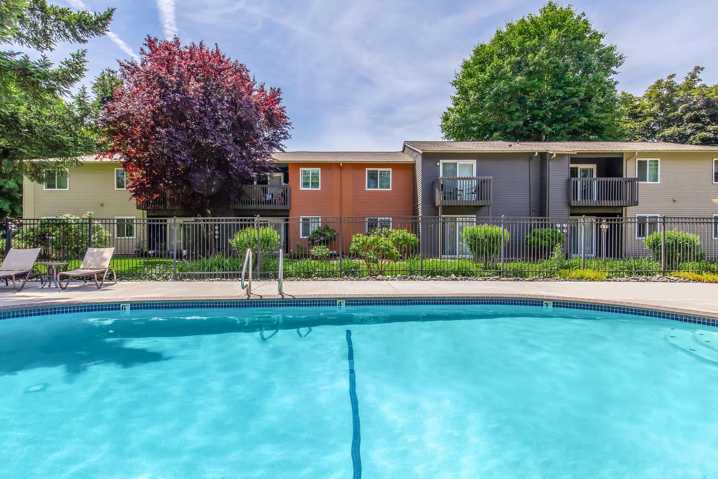 A clear blue swimming pool in the foreground, with lounge chairs along the edge. In the background, there are three apartment buildings with various colors, surrounded by greenery and trees under a bright blue sky. The scene is inviting and peaceful, perfect for relaxation.