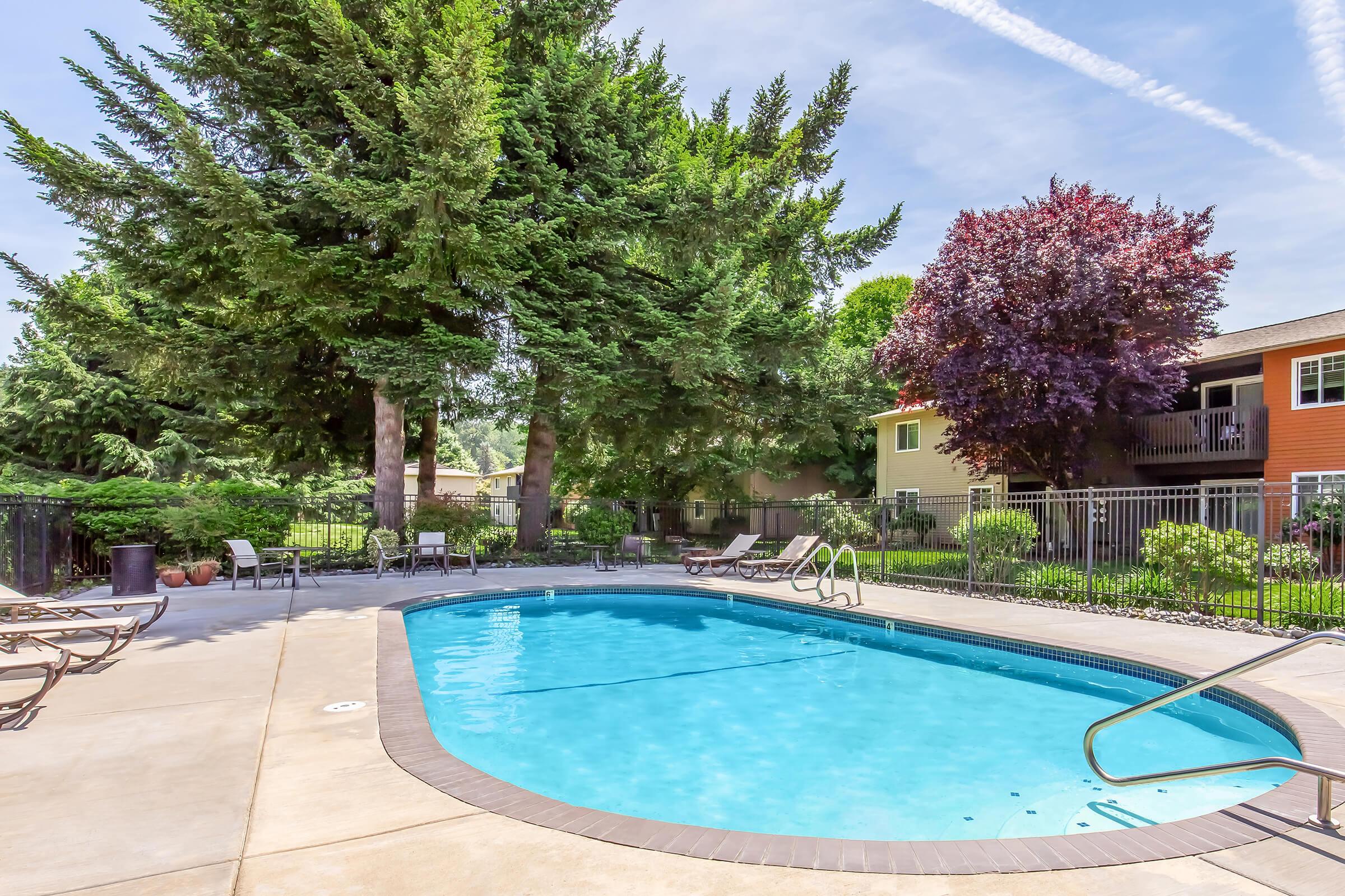A clear blue swimming pool surrounded by trees and lounge chairs. In the background, there are multi-story apartment buildings with colorful facades. The setting is bright and sunny, with blue skies and fluffy clouds, creating a relaxing outdoor atmosphere.