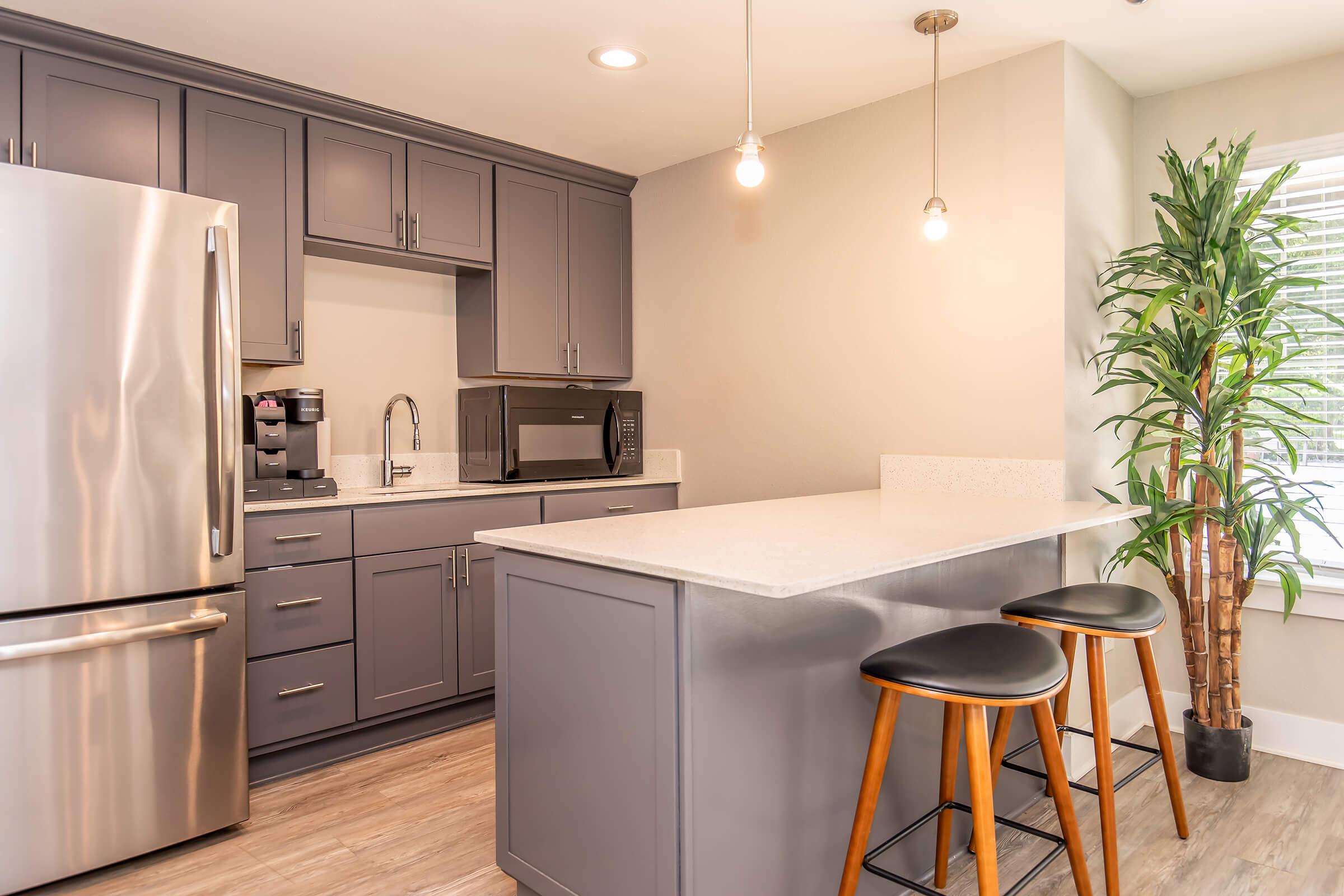 Modern kitchen with gray cabinets, stainless steel refrigerator, and microwave. A white countertop bar with two black stools. A large window with natural light and a potted plant adds a touch of greenery to the space. Ceiling lights provide warm illumination.