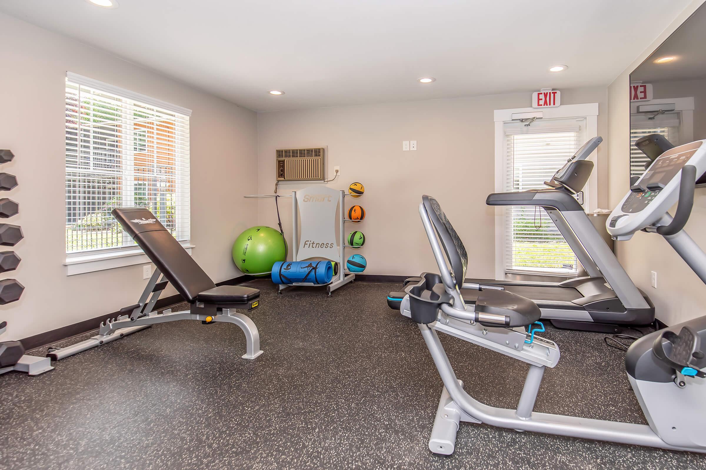 A modern gym interior featuring exercise equipment including a treadmill, stationary bike, and a weight bench. The walls are light-colored with large windows allowing natural light. There's a fitness ball and weights organized neatly in the space, creating a clean and inviting workout environment.
