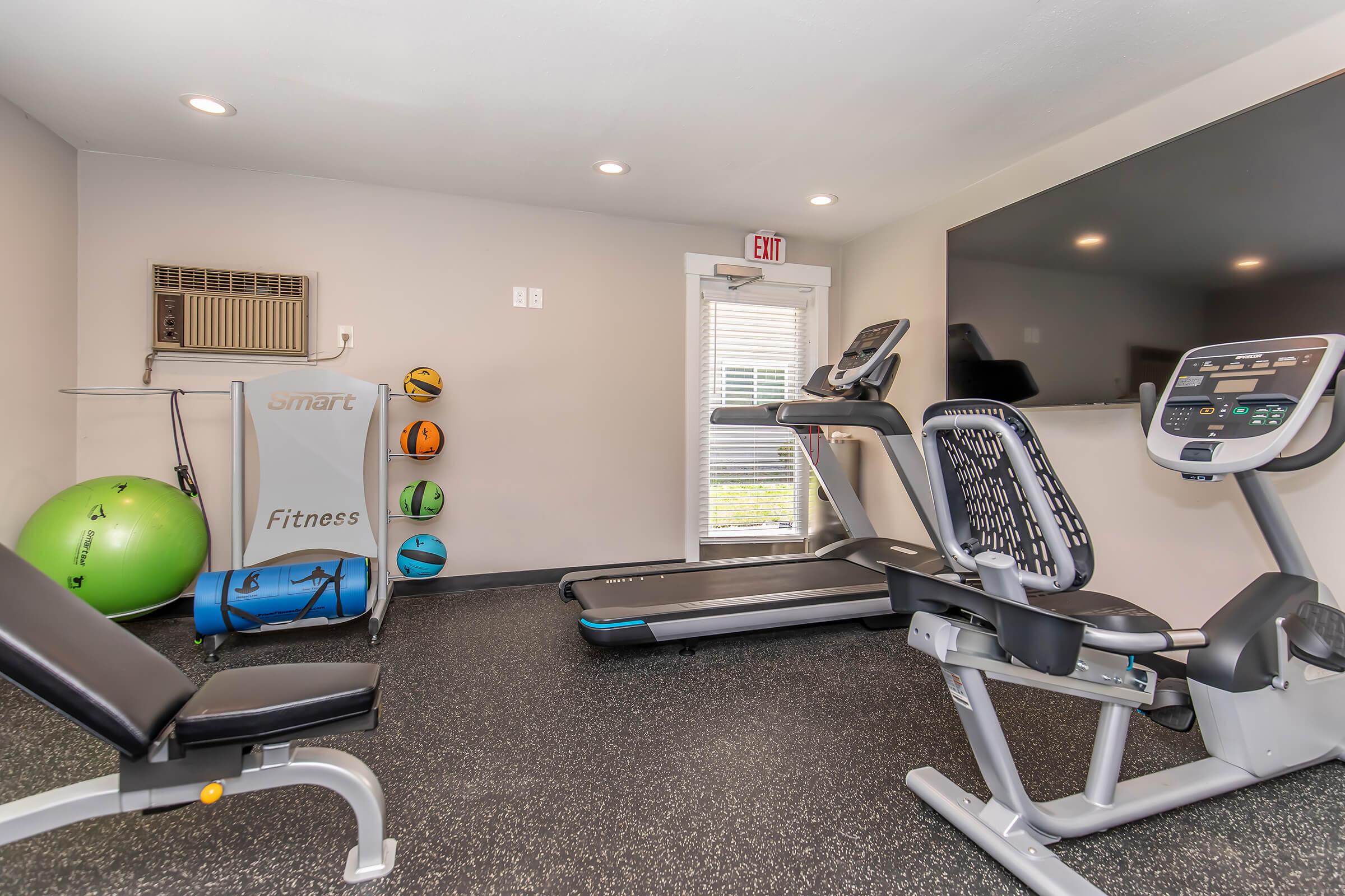 A small fitness room featuring exercise equipment including a treadmill, stationary bike, and a weight bench. The walls are painted light gray, and there's an air conditioning unit as well as colorful exercise balls stored on the side. A large wall-mounted TV is also visible.