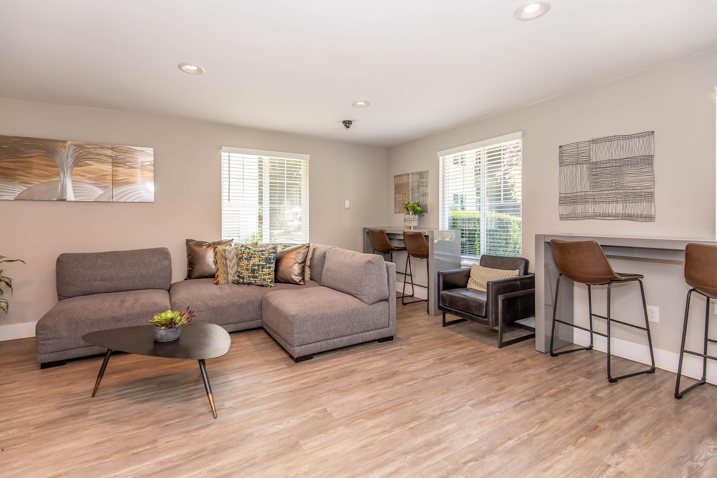 A modern living room featuring a gray sectional sofa with decorative pillows, a black coffee table, and wood-like flooring. Large windows provide natural light, and there are two small bar-height tables with high stools. Wall art adds a contemporary touch to the space.
