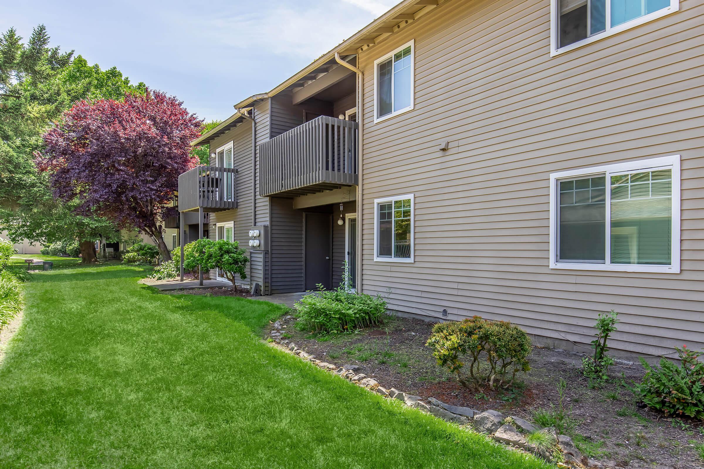 Two-story apartment building with multiple units, featuring large windows and balconies. In the foreground, a green lawn with trimmed bushes and a stone border leads up to the building. Trees are visible in the background, adding a vibrant touch to the landscape.
