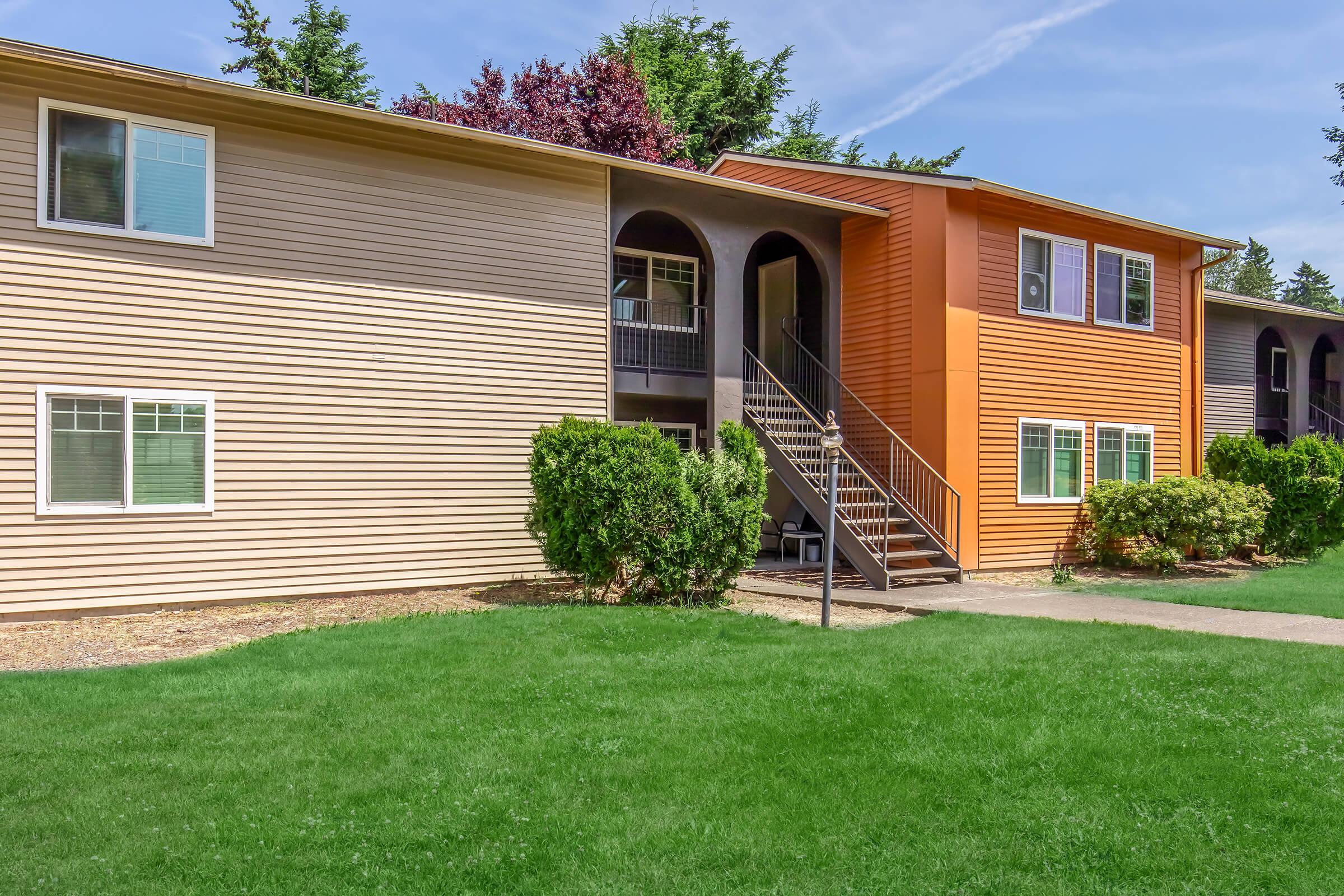 Two multi-unit apartment buildings with a mix of beige and orange siding, surrounded by green grass and small shrubs. The building on the right features a staircase leading to the upper units, while windows are visible on both buildings. Bright blue sky with a few clouds is also visible.