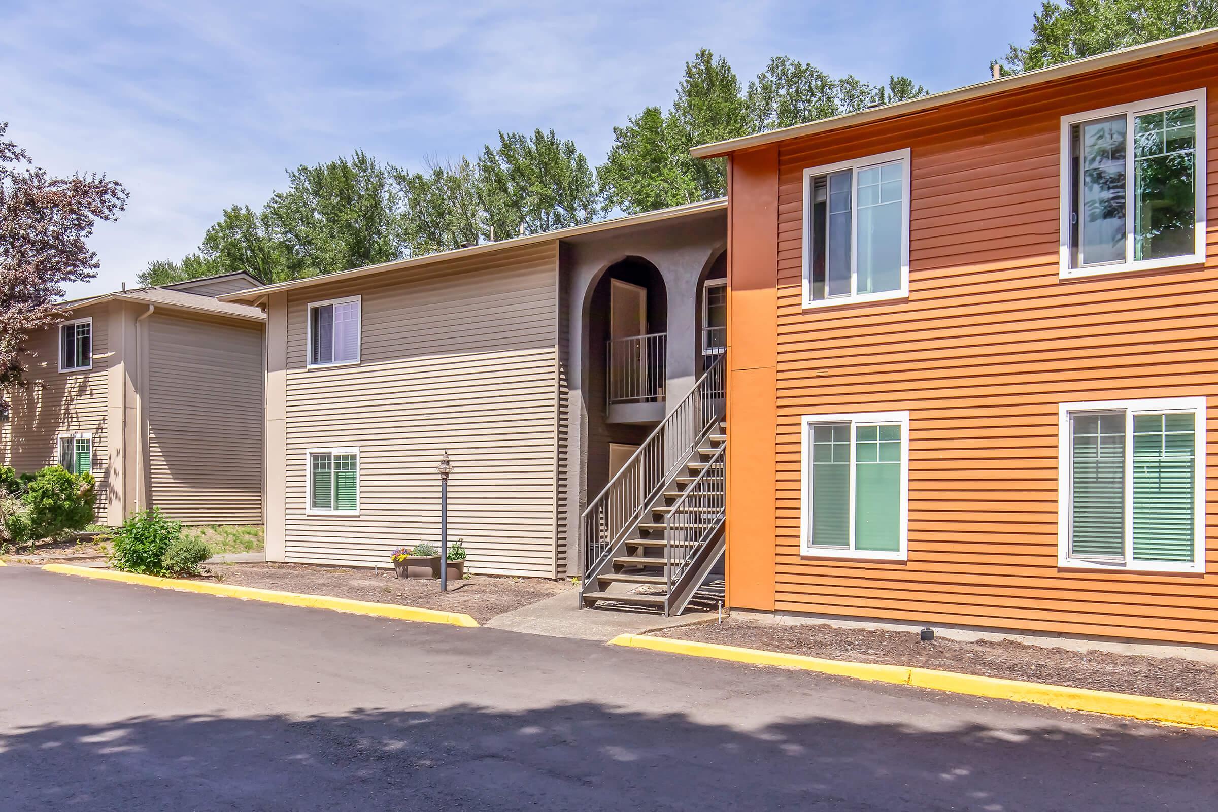 A two-story apartment building with a mix of beige and orange siding. The building features large windows, a staircase leading to the second floor, and a small landscaped area with plants near the entrance. The surroundings include trees and a clear blue sky.