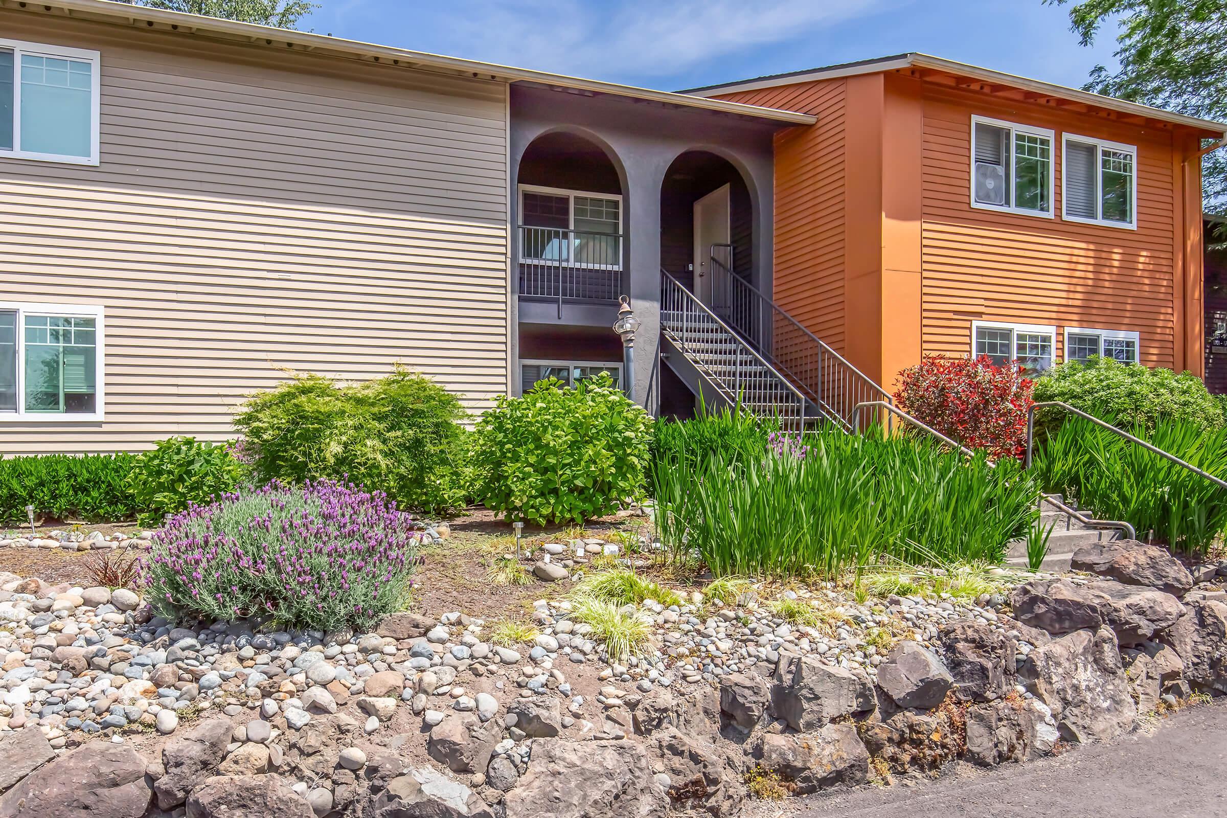 A well-maintained two-story apartment building featuring beige and orange exteriors, surrounded by lush greenery, including flowering plants and shrubs. A stone path leads to the entrance, which has a set of stairs and an arched porch. Clear blue sky overhead enhances the inviting atmosphere.