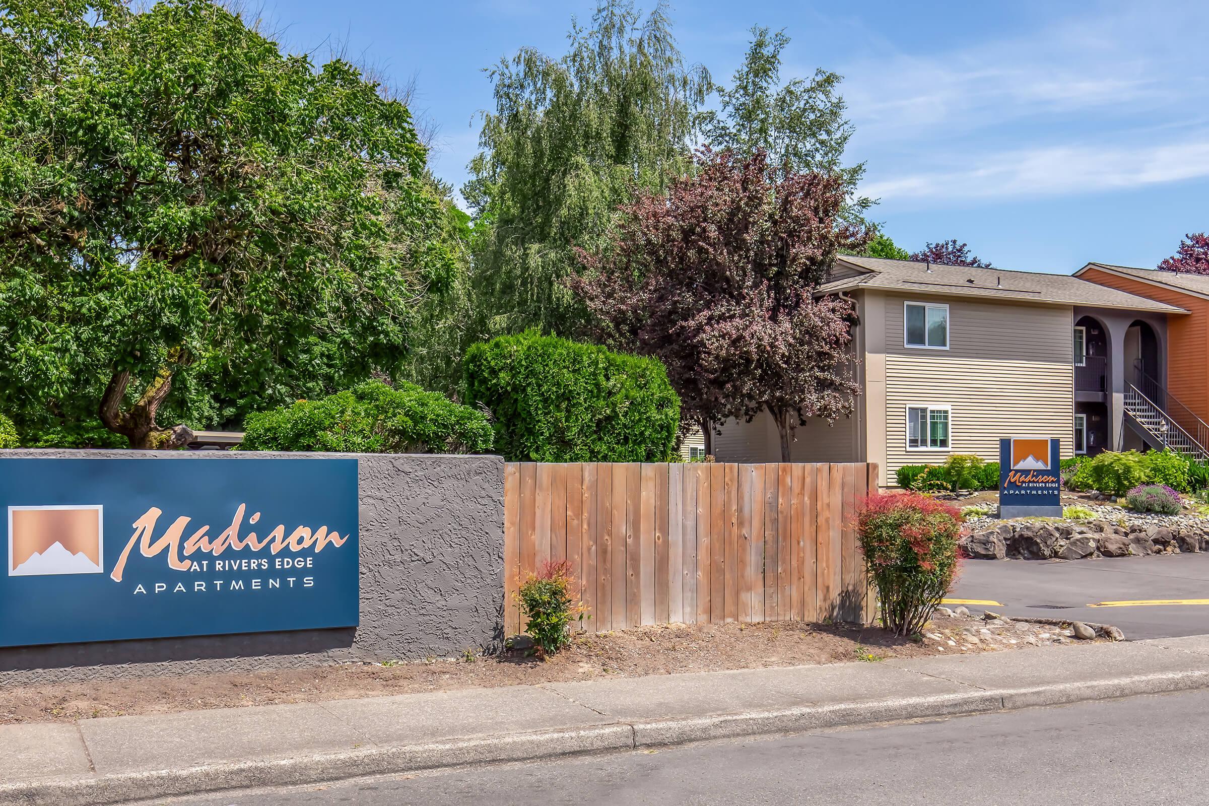 Exterior view of Madison at Rivers Edge Apartments, featuring a prominent sign with the name and logo. Surrounding landscaping includes trees and shrubs, along with a multi-unit residential building in the background. The setting appears well-maintained and inviting.