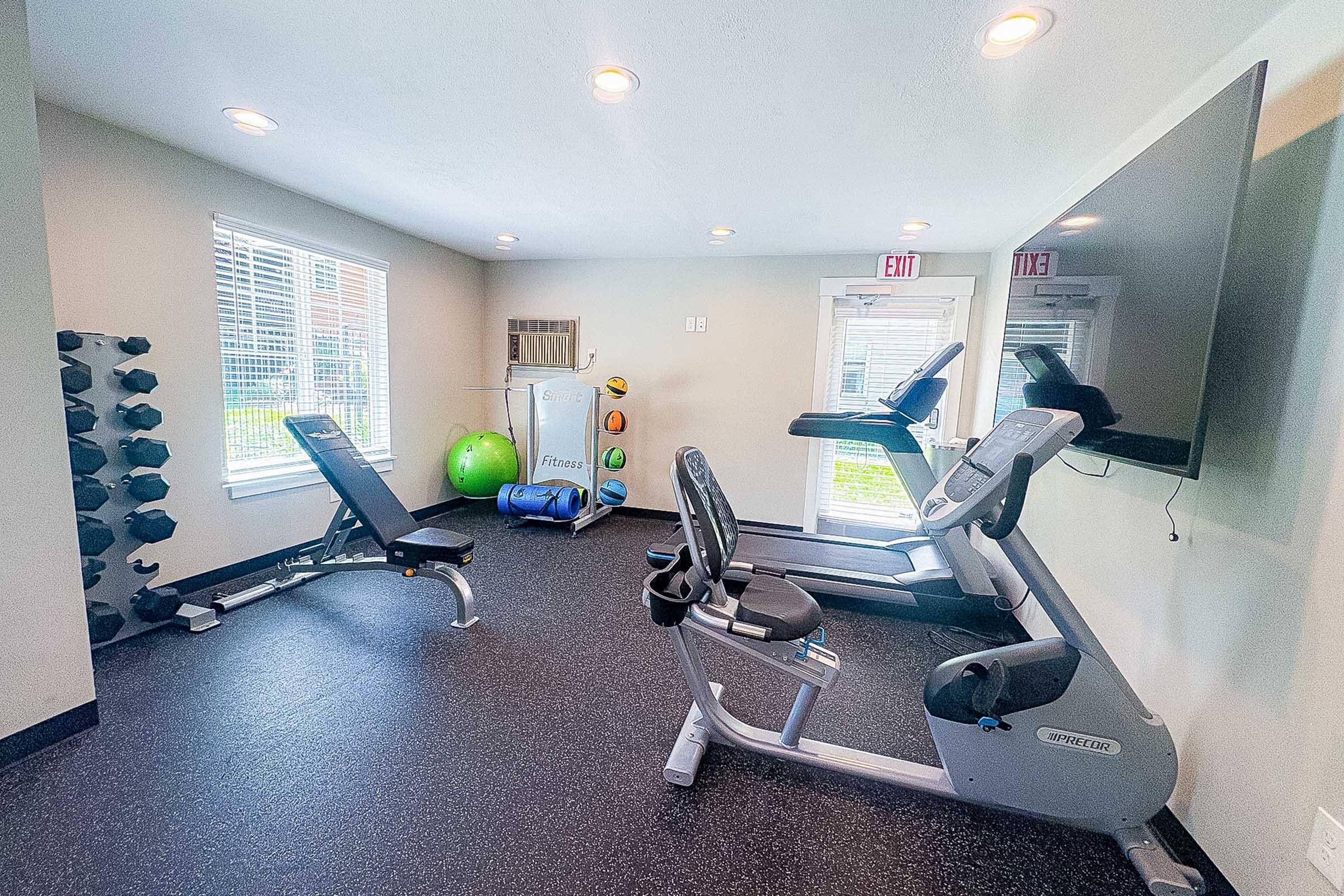 A small gym room featuring exercise equipment, including a stationary bike, a weight bench, and free weights. There's a large TV mounted on the wall and large windows allowing natural light. The floor is covered with black rubber flooring, and exercise balls are visible in the corner.