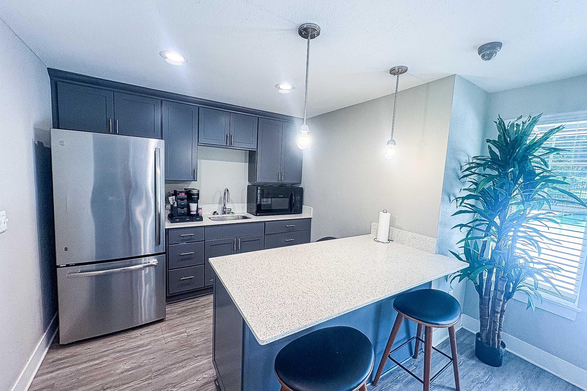 Modern kitchen featuring a stainless steel refrigerator, microwave, and coffee maker. Gray cabinetry complements a light countertop. A small island with two wooden bar stools sits in the center, and a tall plant adds greenery. Bright lighting from overhead fixtures enhances the open, inviting space.