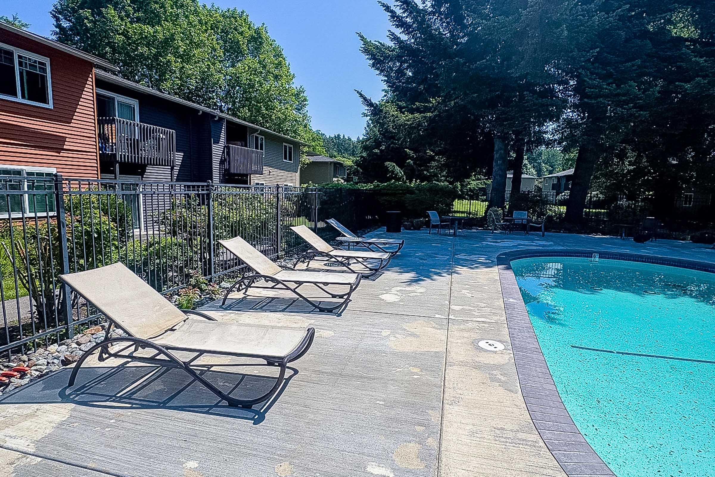 A sunny poolside scene featuring a clear blue pool with lounge chairs arranged neatly around it. Lush greenery and trees are visible in the background, along with two multi-story buildings. The area is well-maintained, creating a relaxing outdoor atmosphere.